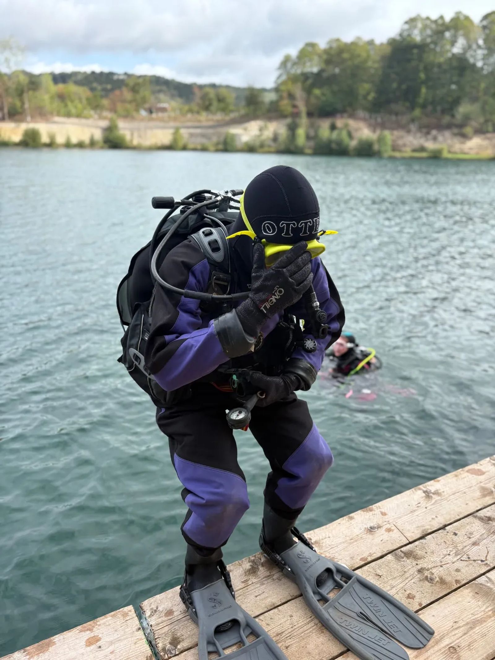 A diver in a dry suit and fins standing on the jetty, hand over the mask, about to enter the water