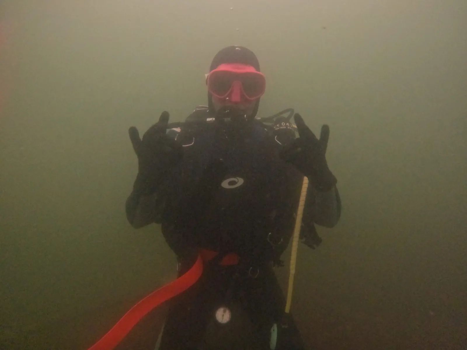 A diver in the green murk of a flooded gravel pit, throwing rock horns at the camera