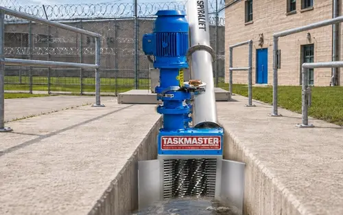 TASKMASTER grinder with SPIRALIFT screw conveyor installed in a concrete wastewater channel outside a correctional facility with razor-wire fencing