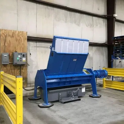 TASKMASTER XL pallet shredder installed inside a plant, with its closed-top single pallet hopper raised, yellow guard rails around the machine, a wall-mounted control panel behind it, and a grey forklift waste hopper positioned under the discharge