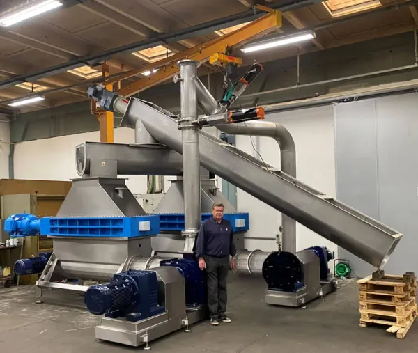 A man standing in a warehouse next to a large industrial, dual shredder machine with a conveyor attachment