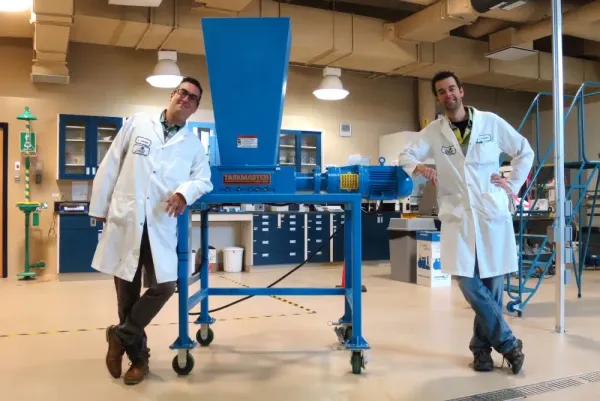 Two men in lab coats standing next to an industrial shredder in a warehouse