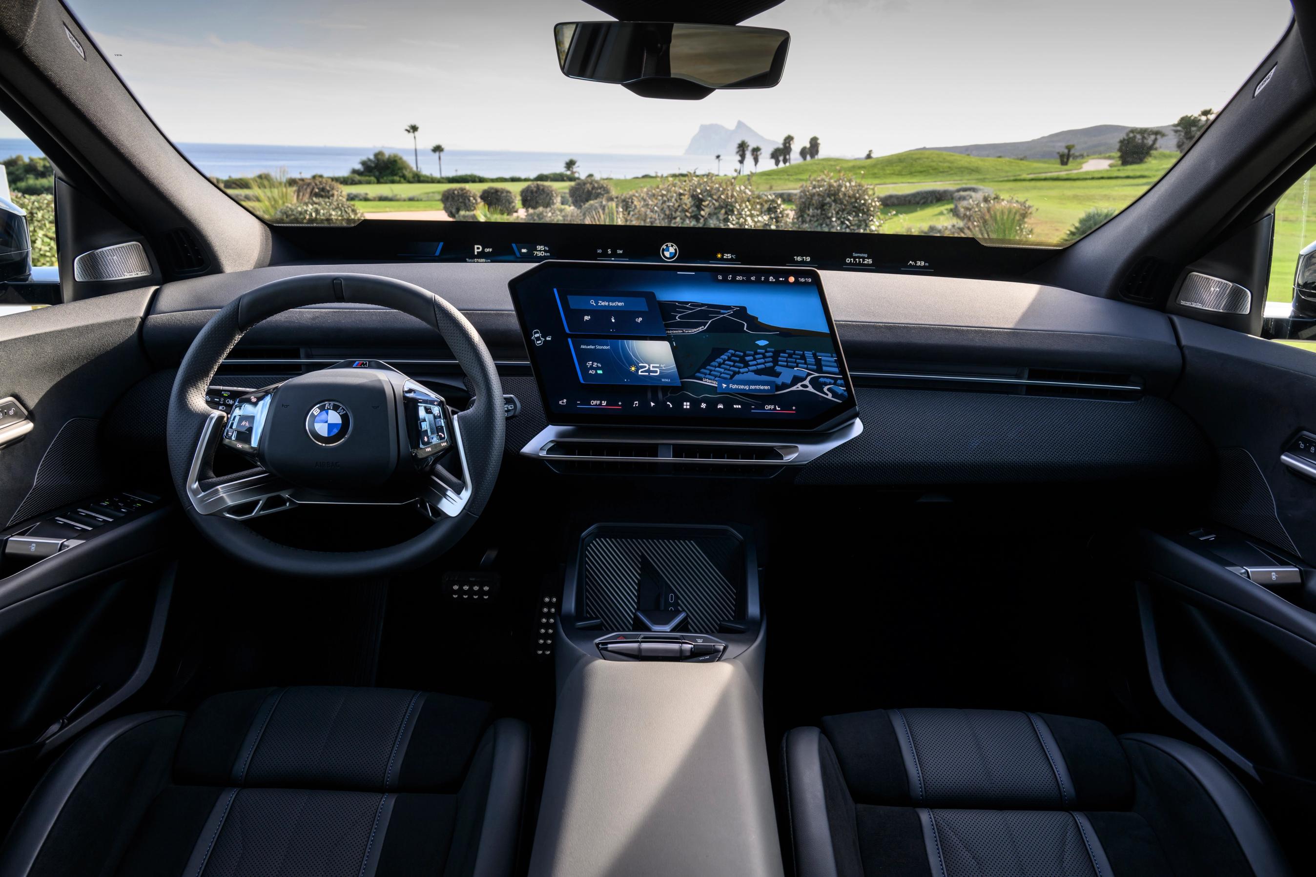 Førerpladsen i en BMW iX3 med græsmark og hav i baggrunden