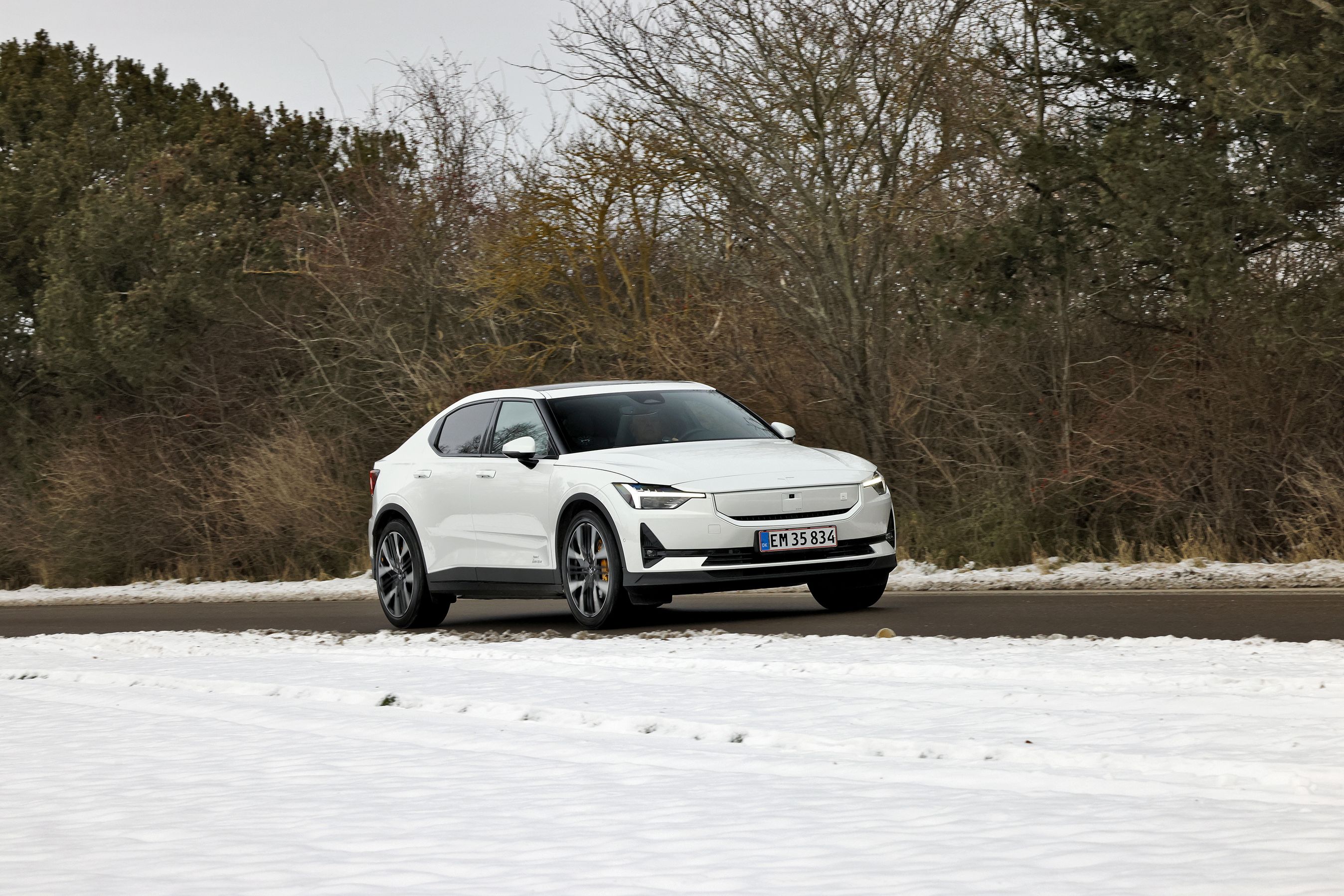 Polestar 2 Performance i hvid farve kører på en landevej med træer i baggrunden og sne i forgrunden.