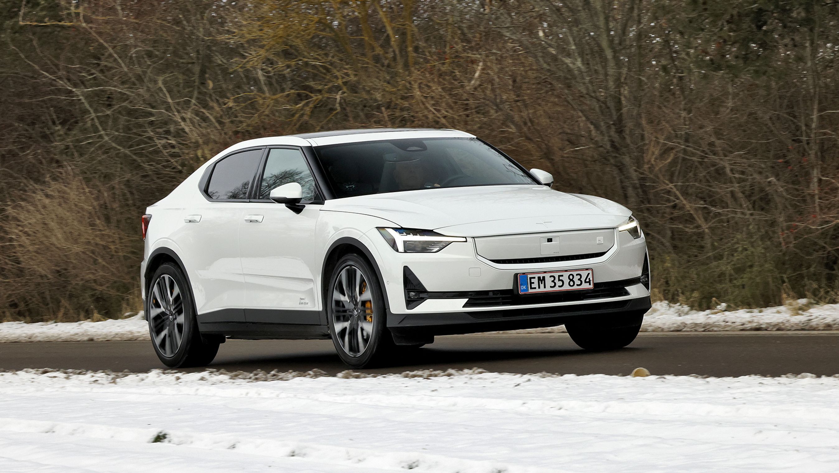 Polestar 2 Performance i hvid farve kører på en landevej med træer i baggrunden og sne i forgrunden.