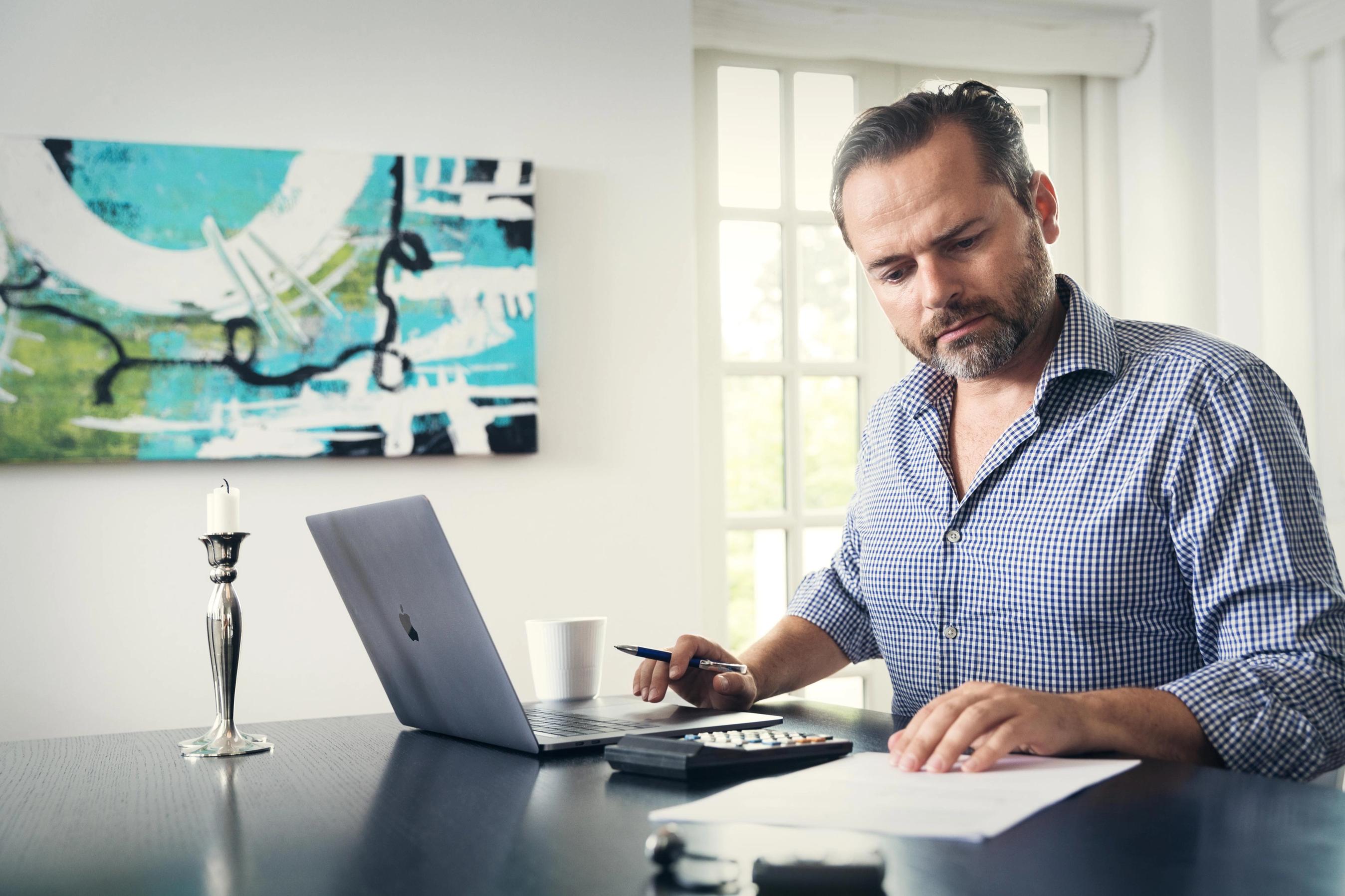 Mand sidder ved bærbar computer indenfor og kigger på papirer.