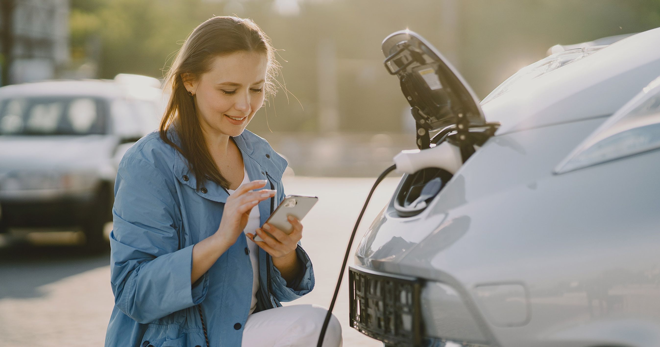kvinde lader sin elbil ved ladestander og kigger på sin mobil. 