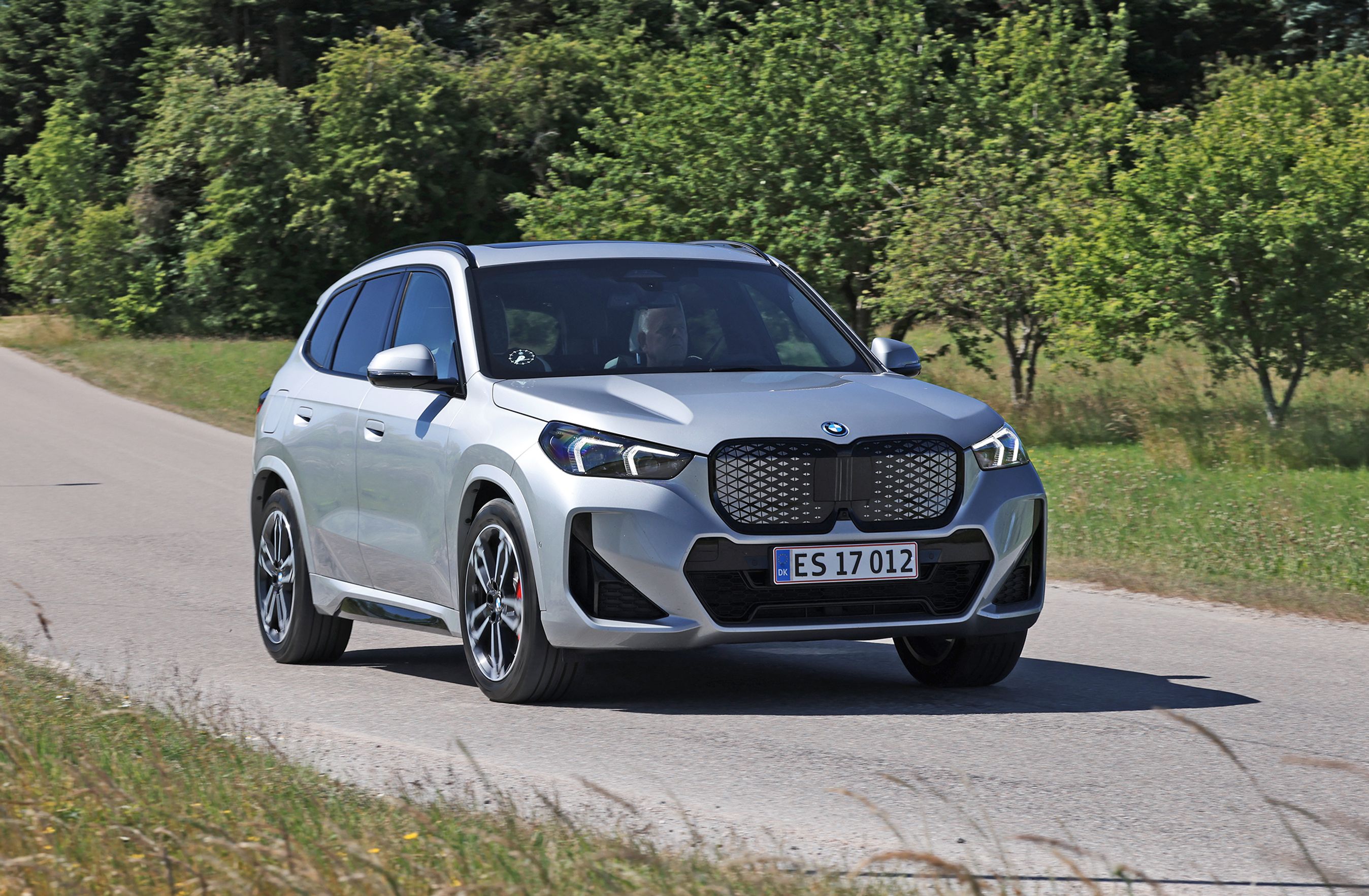 Den faceliftede udgave af BMW iX1 eDrive Supercharged fra 2026 set forfra på landevej med grønne træer i baggrunden.