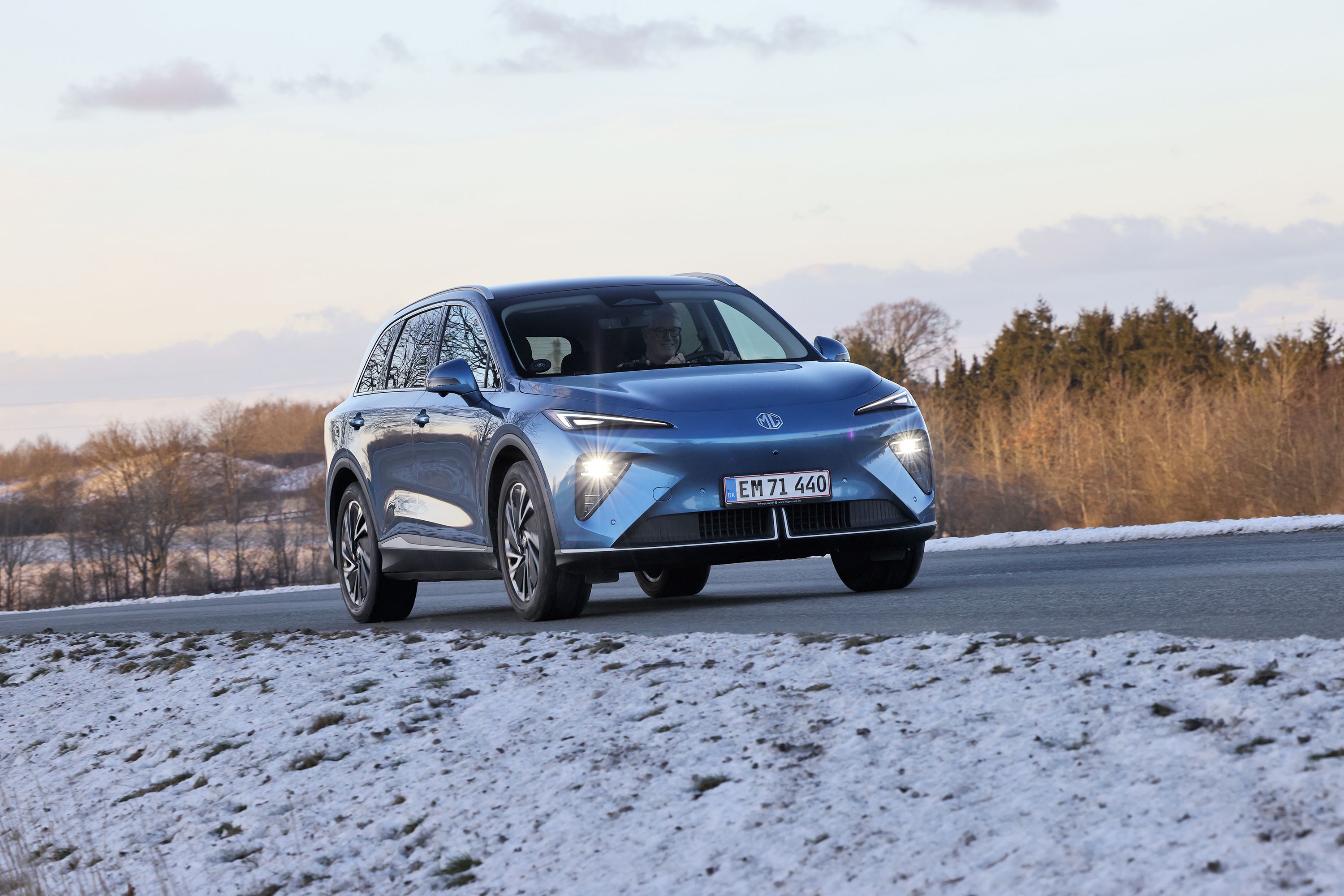 En blå MG MGS6 kører på en landevej med træer i baggrunden og sne på jorden i forgrunden. 