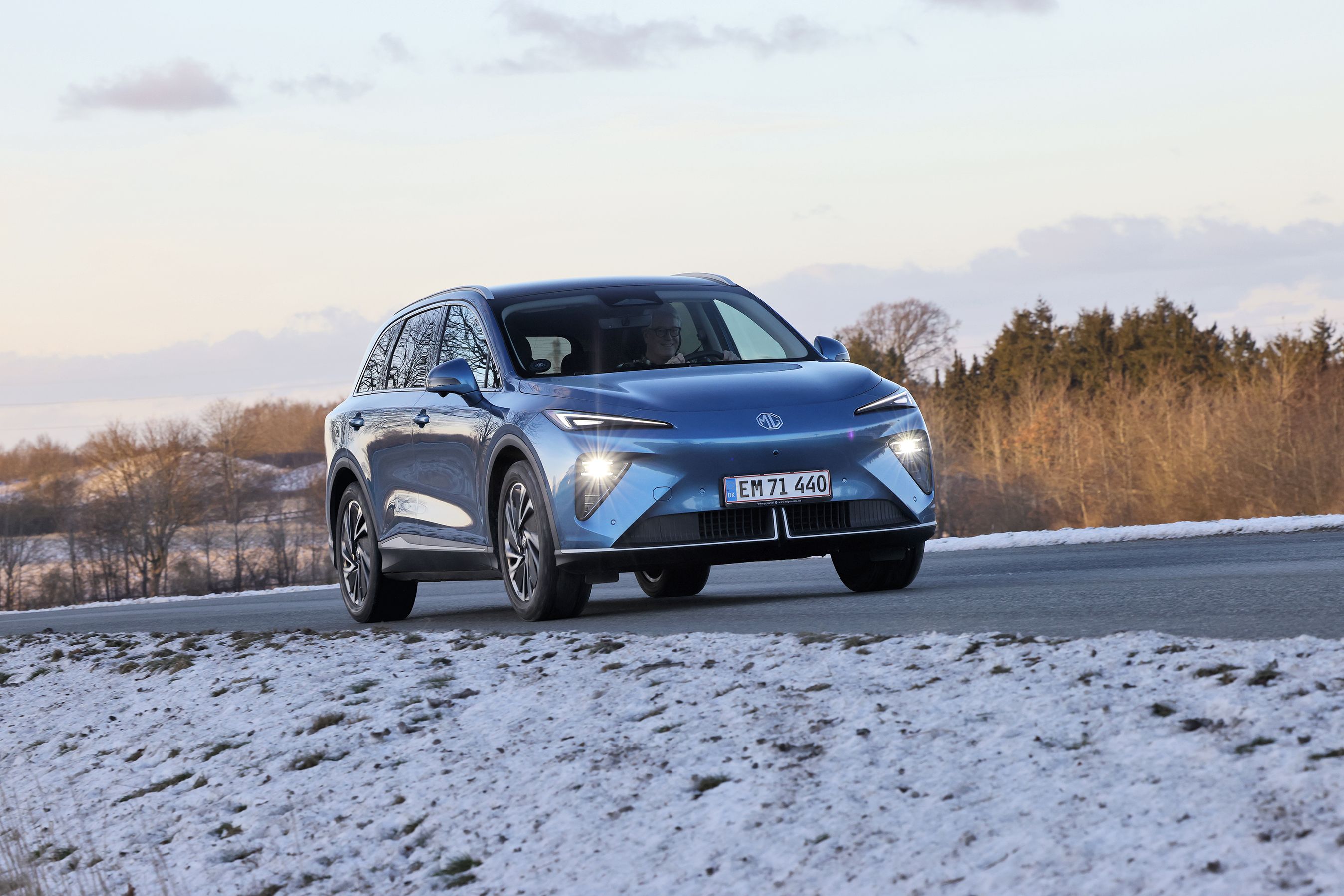En blå MG MGS6 kører på en landevej med træer i baggrunden og sne på jorden i forgrunden.