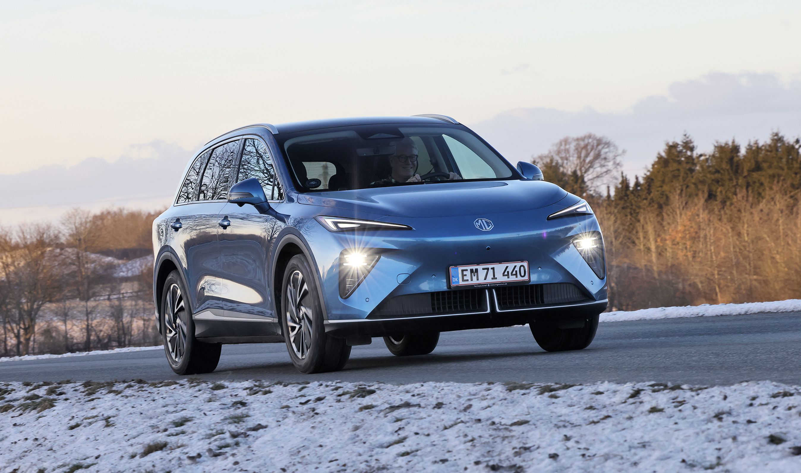 En blå MG MGS6 kører på en landevej med træer i baggrunden og sne på jorden i forgrunden.