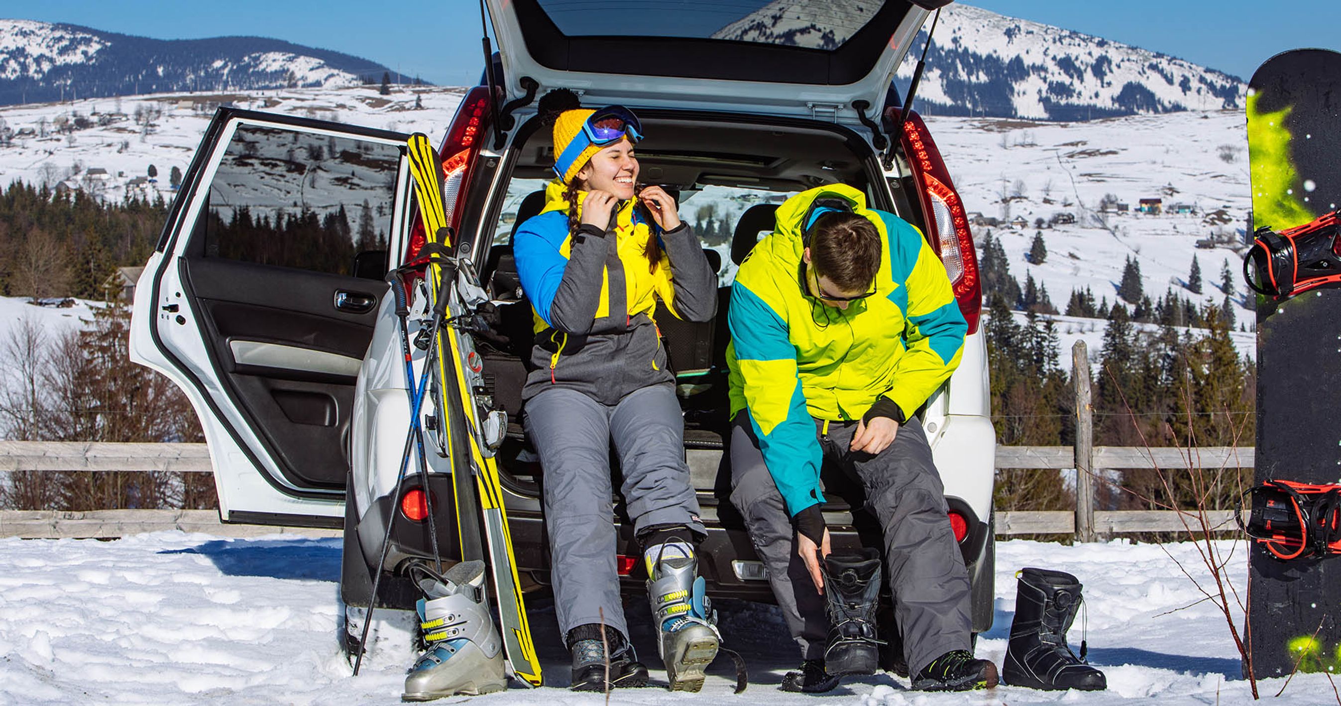 Bil holder foran bjerge, og to personer er ved at tage skitøj og -udstyr på.