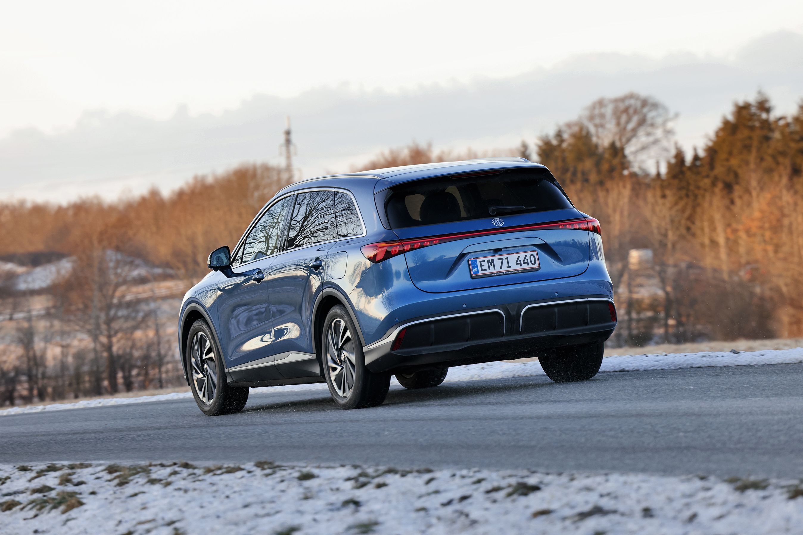 En blå MG MGS6 set bagfra kørende på en vej omgivet af sne og med træer i baggrunden.