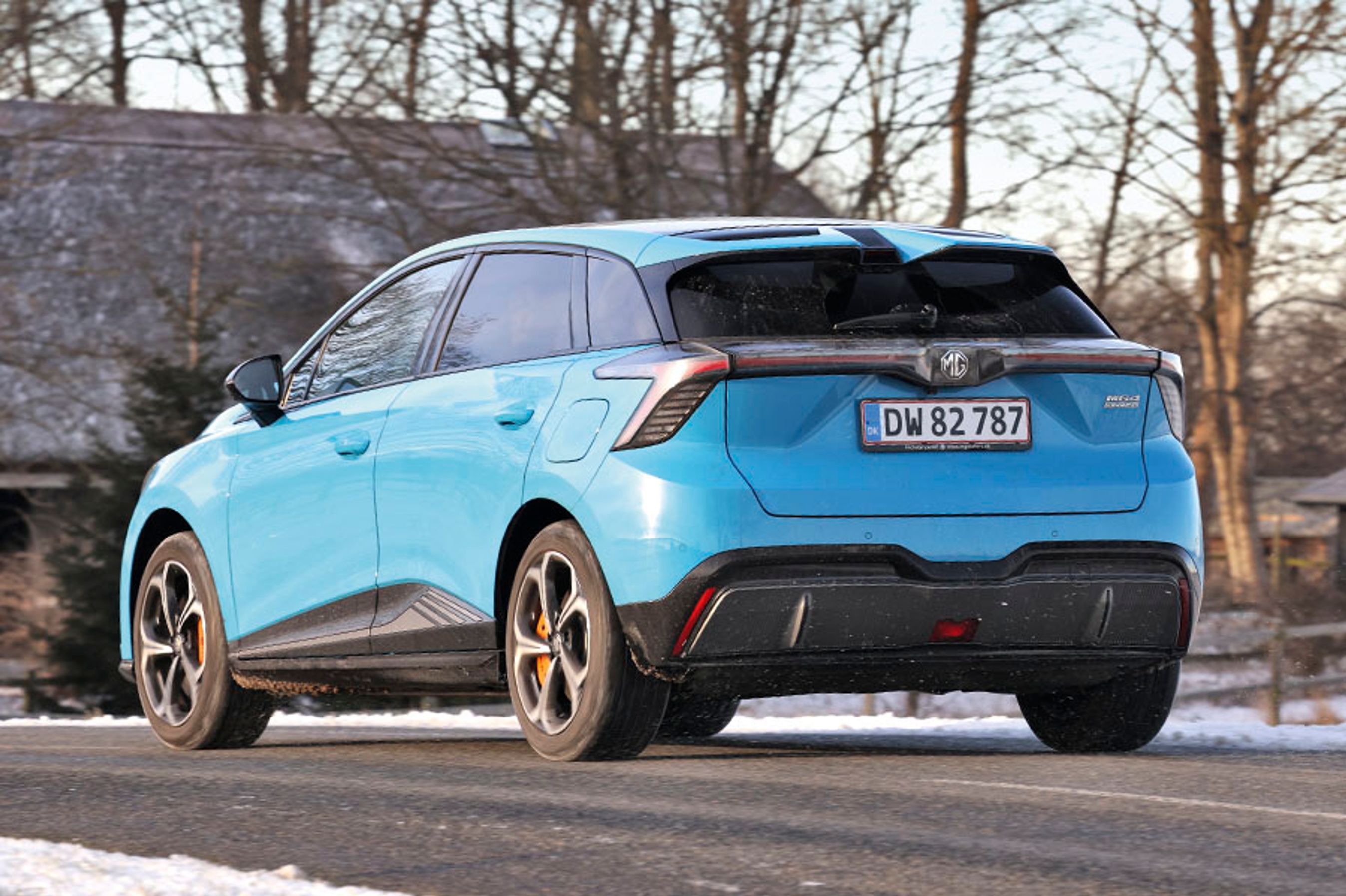 En lyseblå MG 4 XPower set bagfra, mens den kører på en vej med et hus i baggrunden