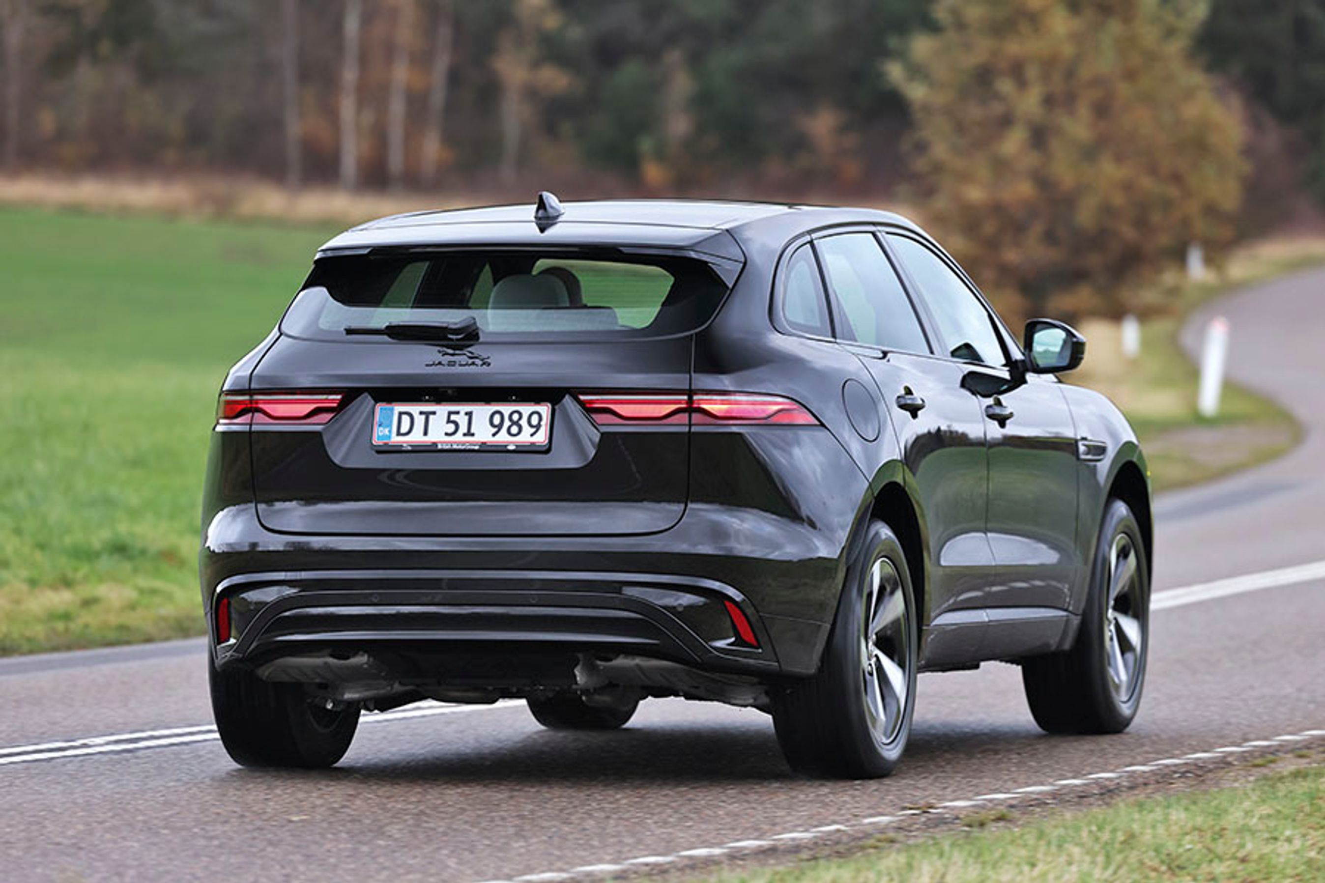 En sort Jaguar F-Pace set bagfra, mens den kører ind i et sving på en landevej med græs og træer i baggrunden