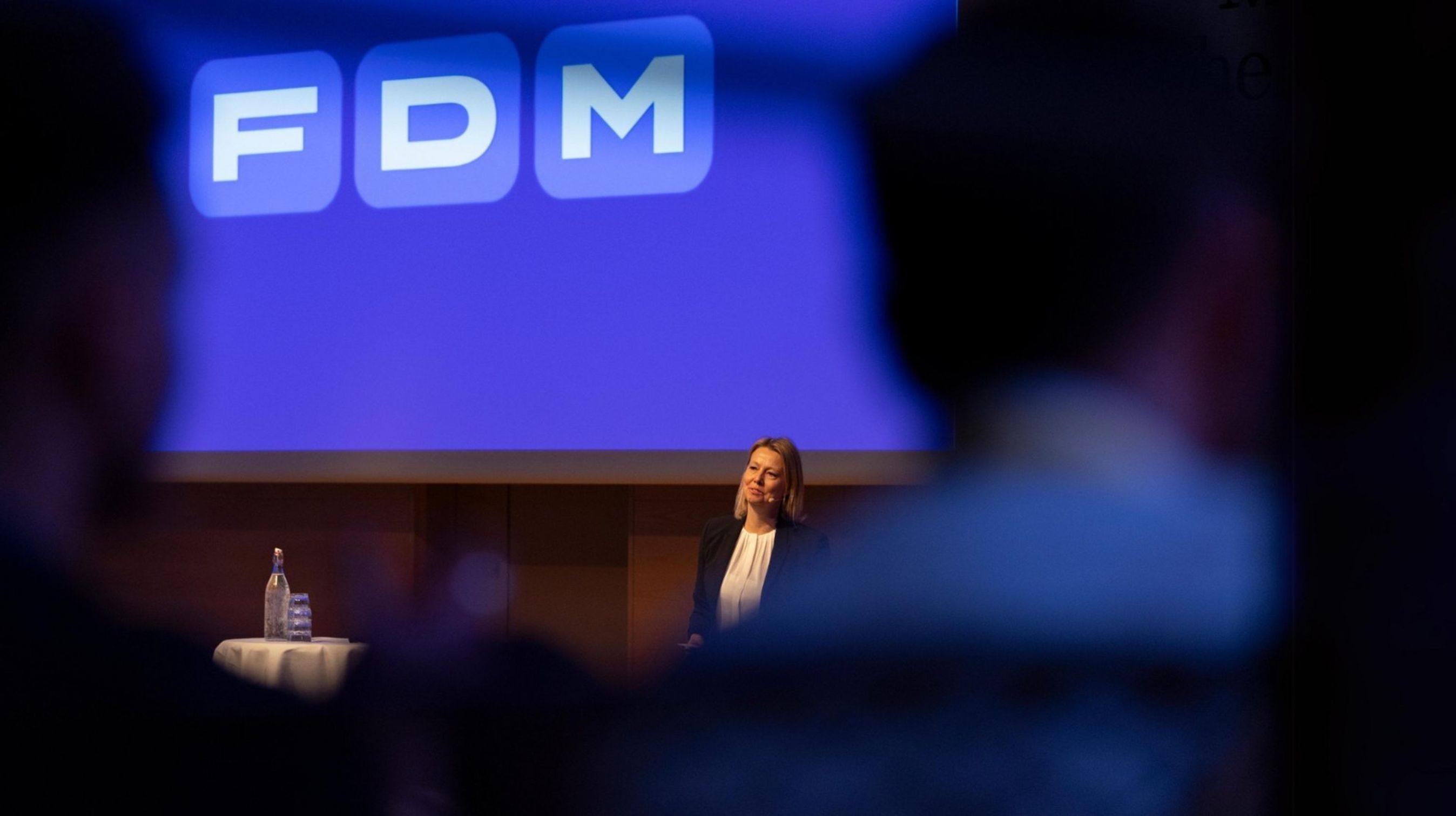 FDMs administrerende direktør, Stina Glavind, holder oplæg foran blå skærm med FDM-logo. Silhouetter af mennesker i forgrunden.