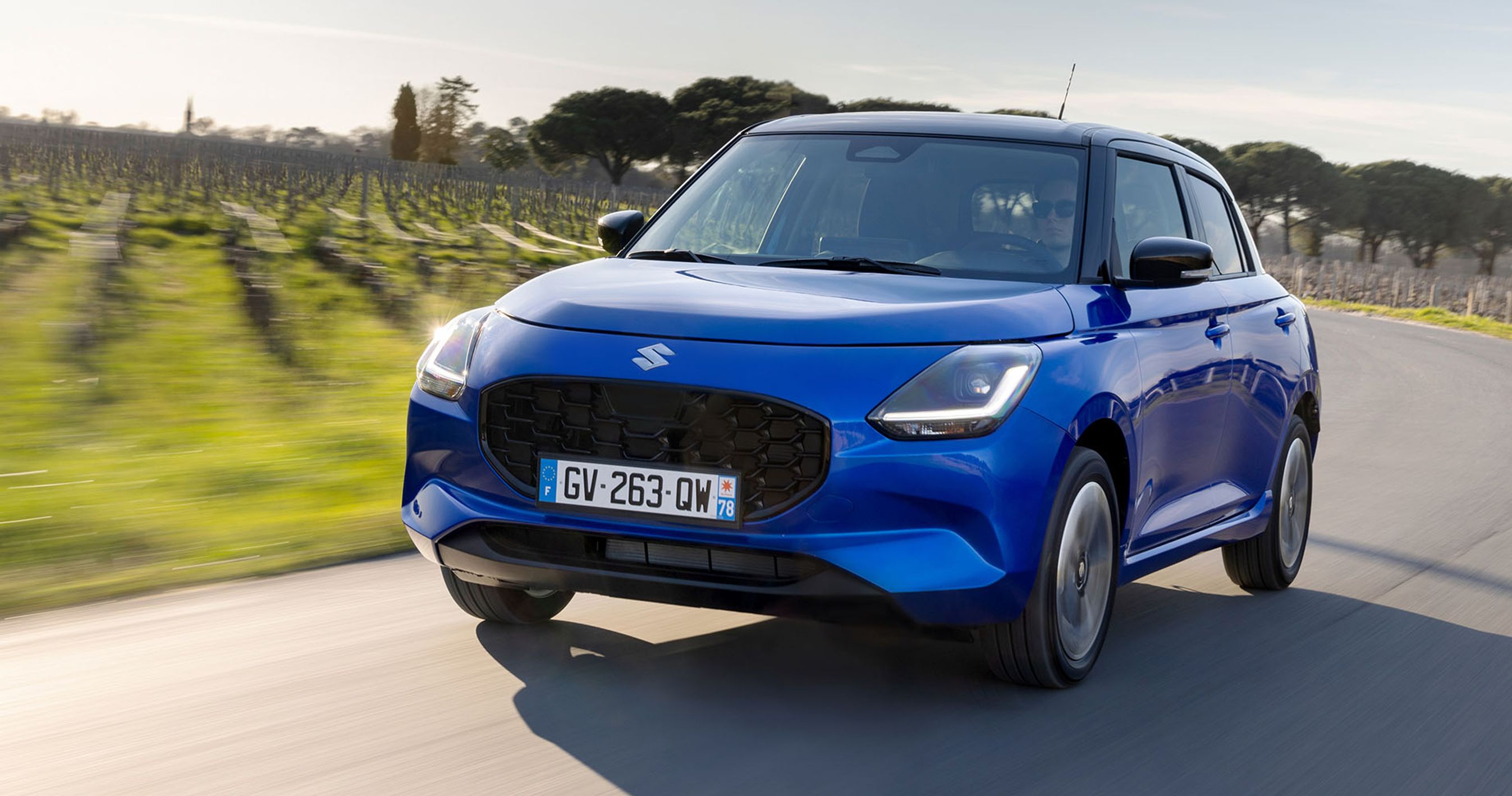 Blå Suzuki Swift kører på en vej med vinmarker i baggrunden