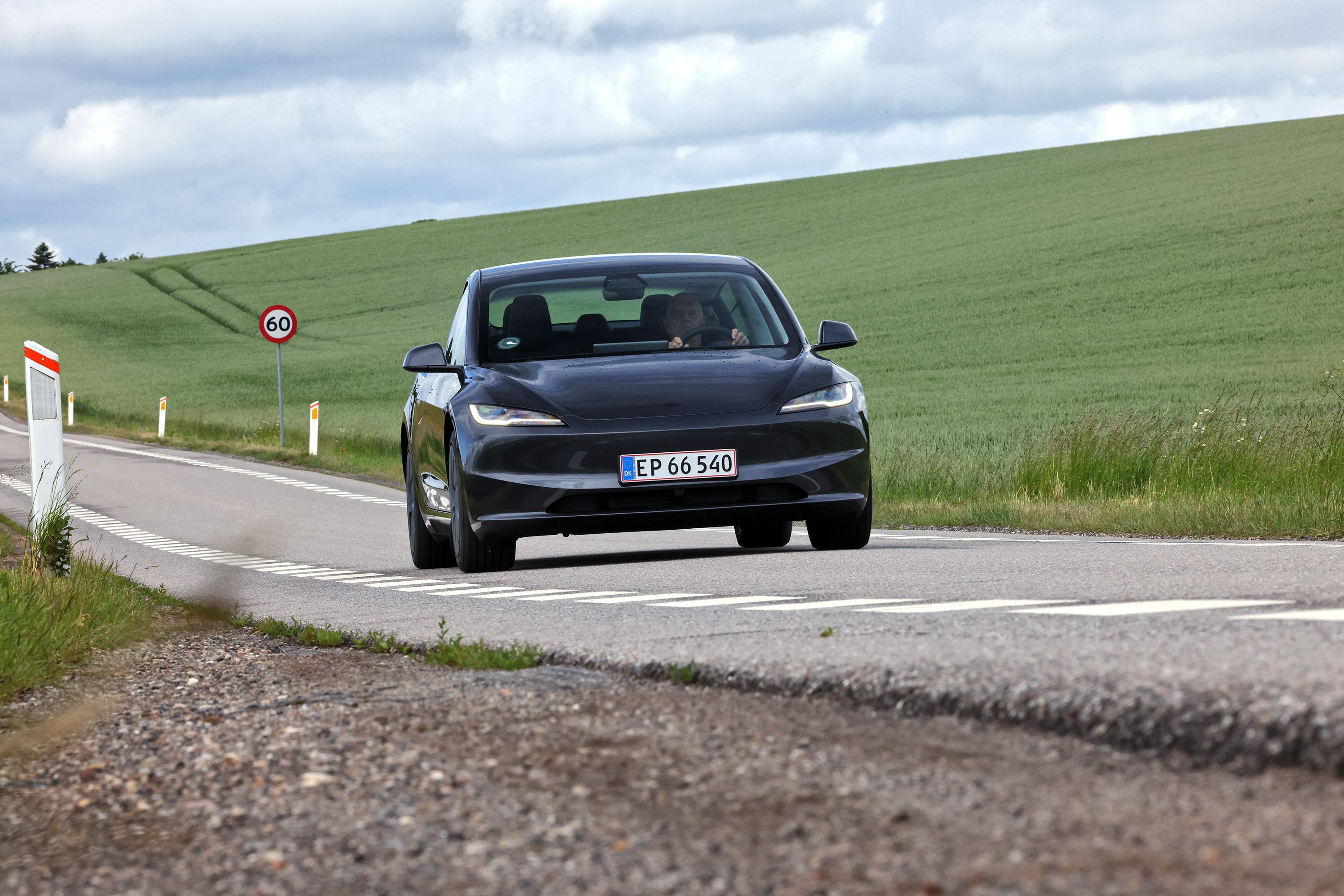  Tesla Model 3 baghjulstræk fra 2026 kører på en dansk landevej med grønne marker i baggrunden og en farttavle med 60 km/t. 