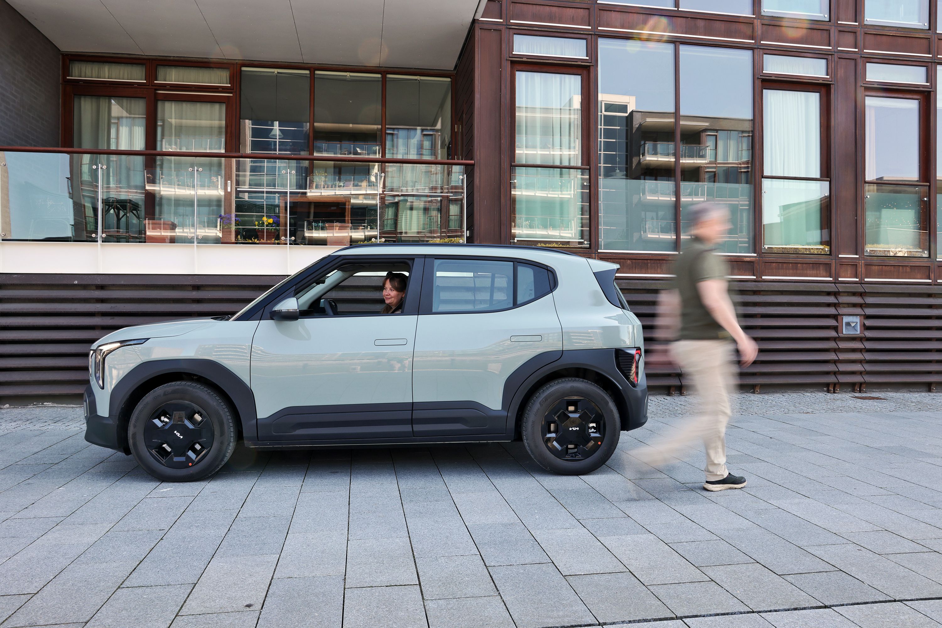 Kia EV2 set fra siden, mens en person sidder i bilen og en anden går forbi