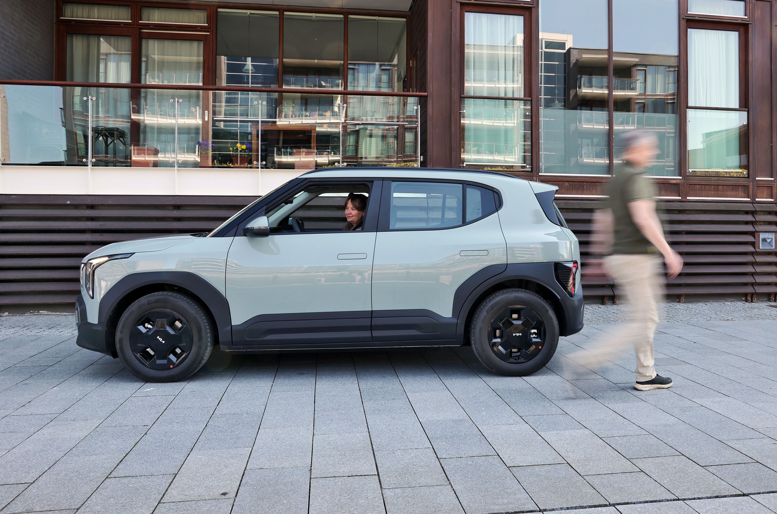 Kia EV2 set fra siden, mens en person sidder i bilen og en anden går forbi