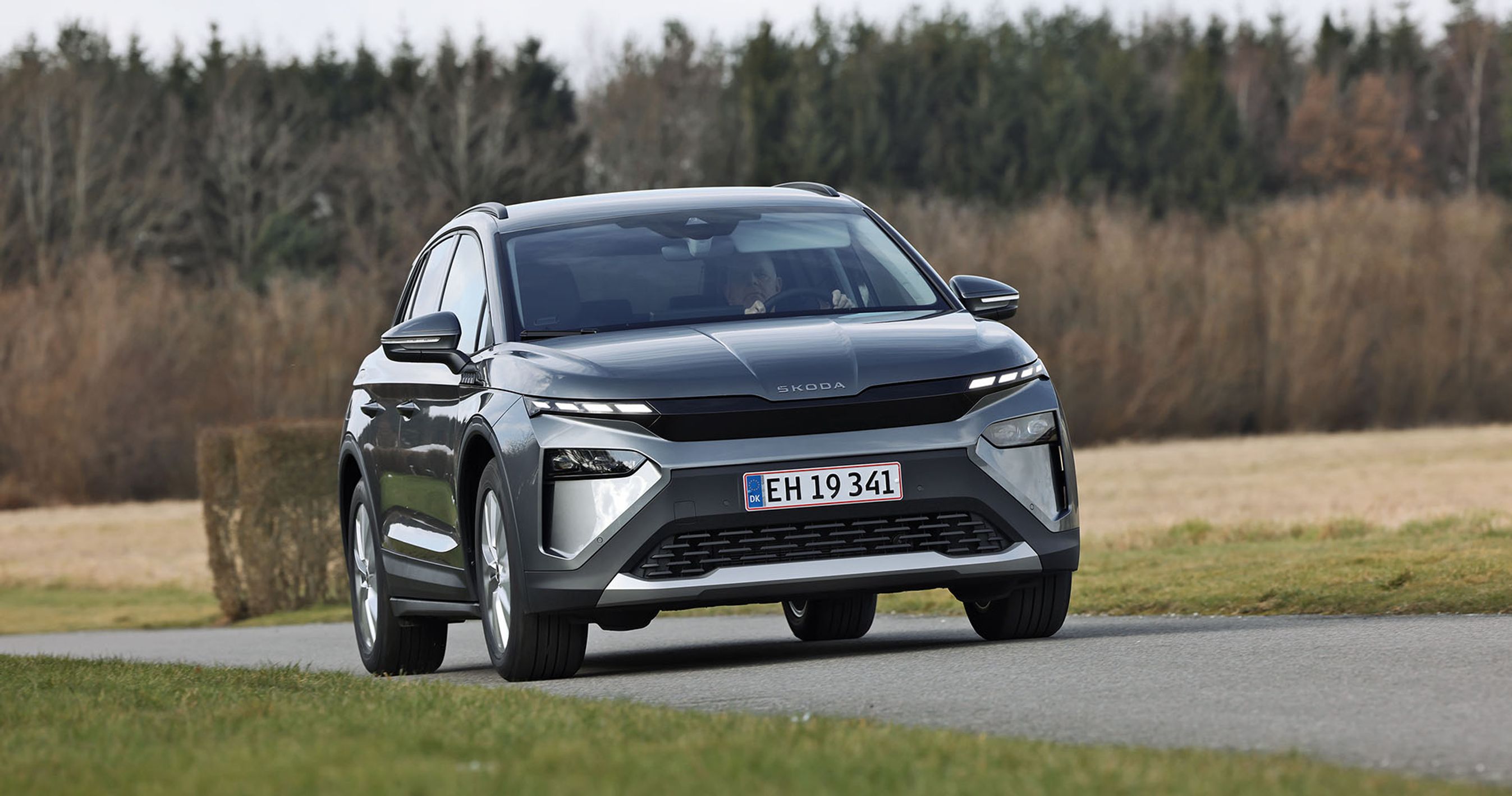 Skoda Elroq 50 set forfra, mens den kører på en landevej med træer og marker i baggrunden
