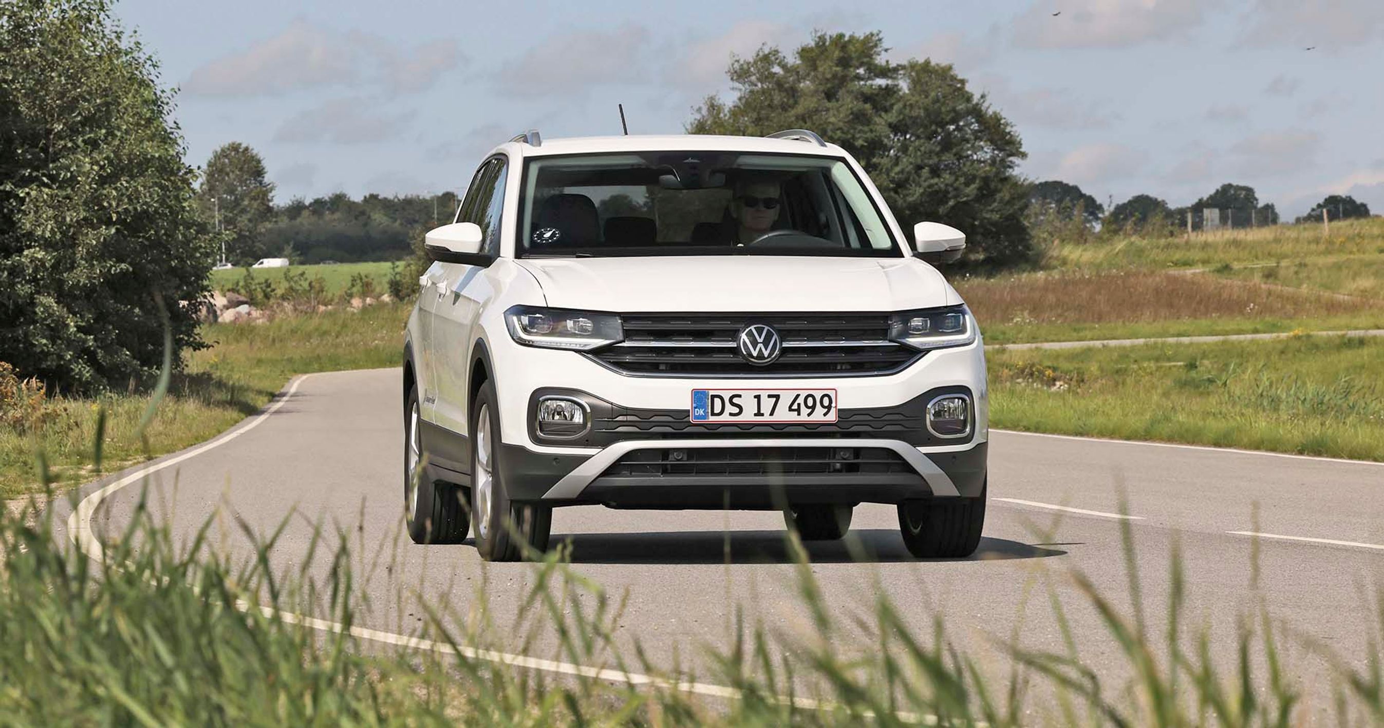 En hvid VW T-Cross mini-SUV kører på en landevej, der svinger. Græs i for- og baggrunden.