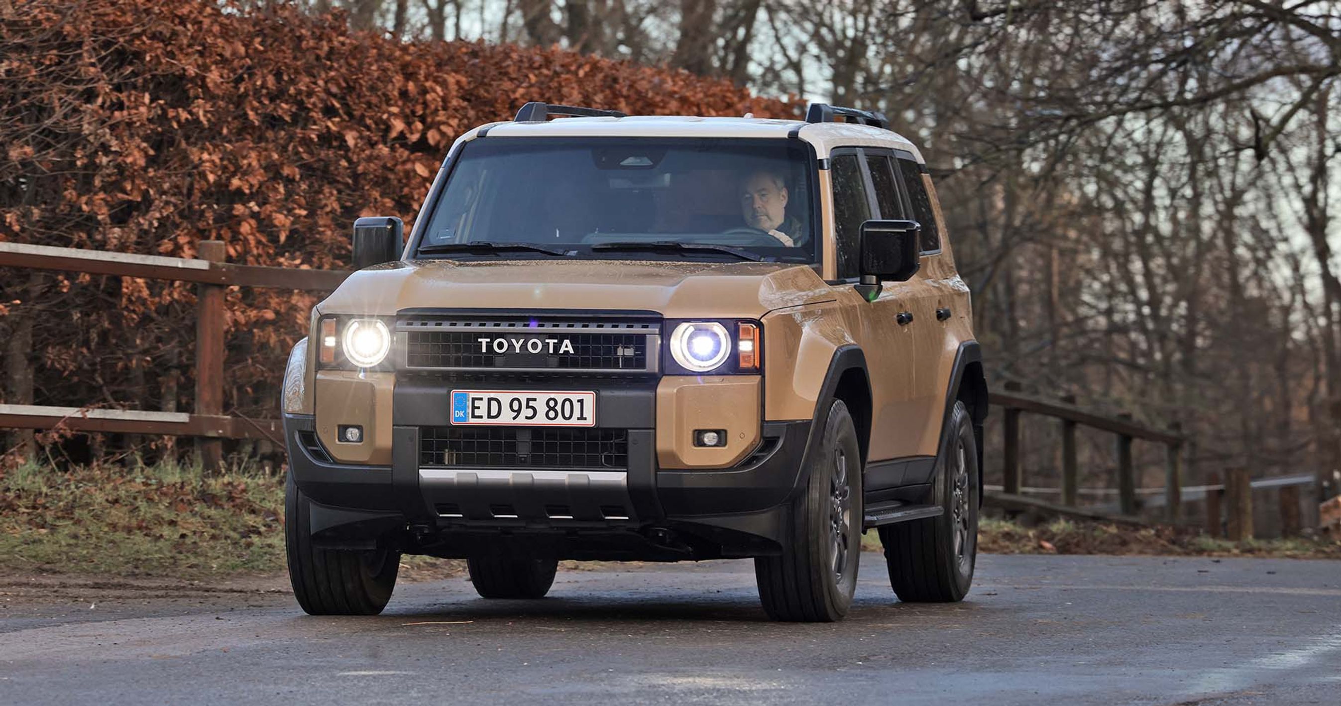 En Toyota Land Cruiser set skråt forfra kører på landevej med hegn og buske i baggrunden