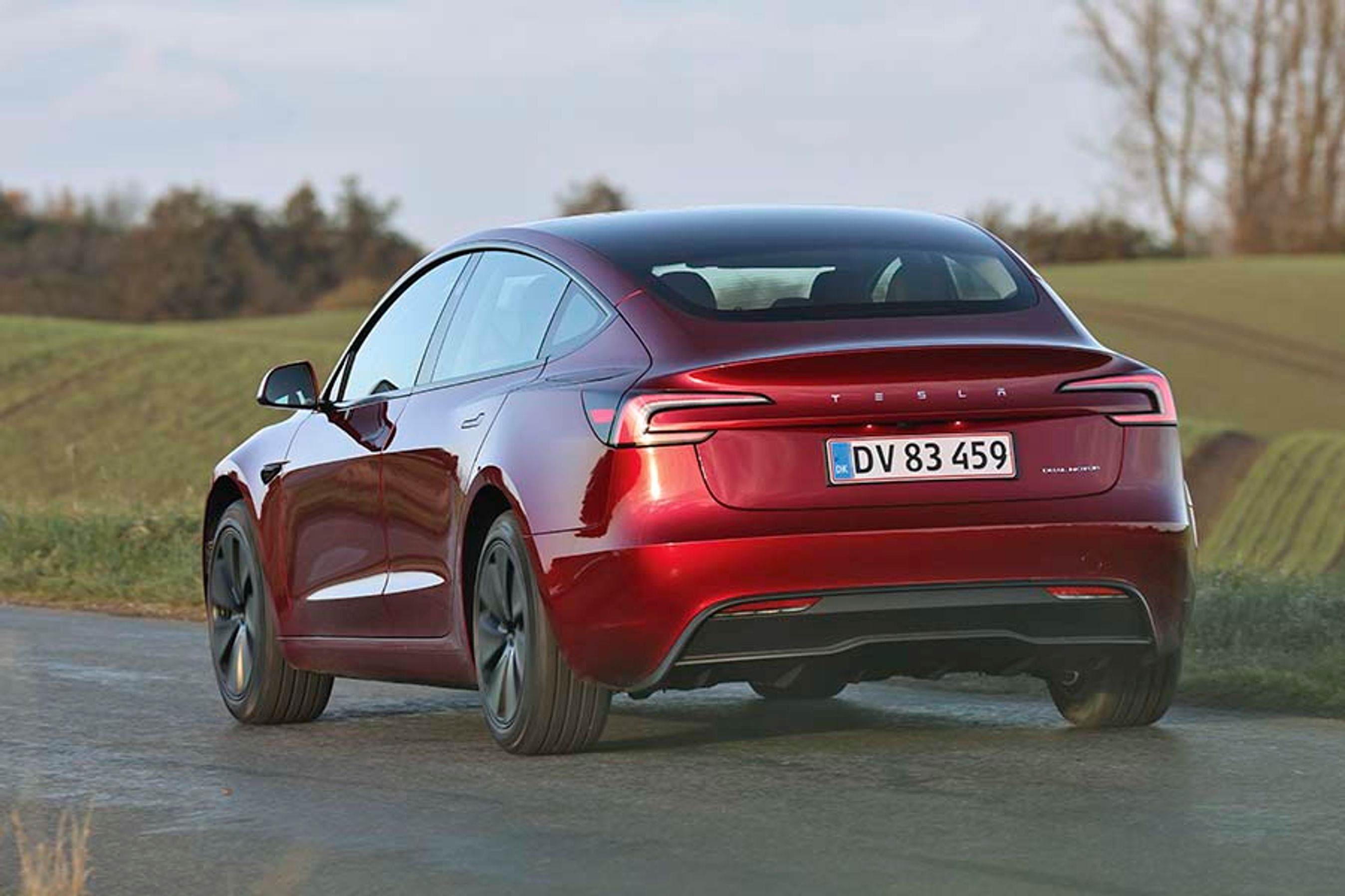 En rød Tesla Model 3 set bagfra, mens den kører på en vej
