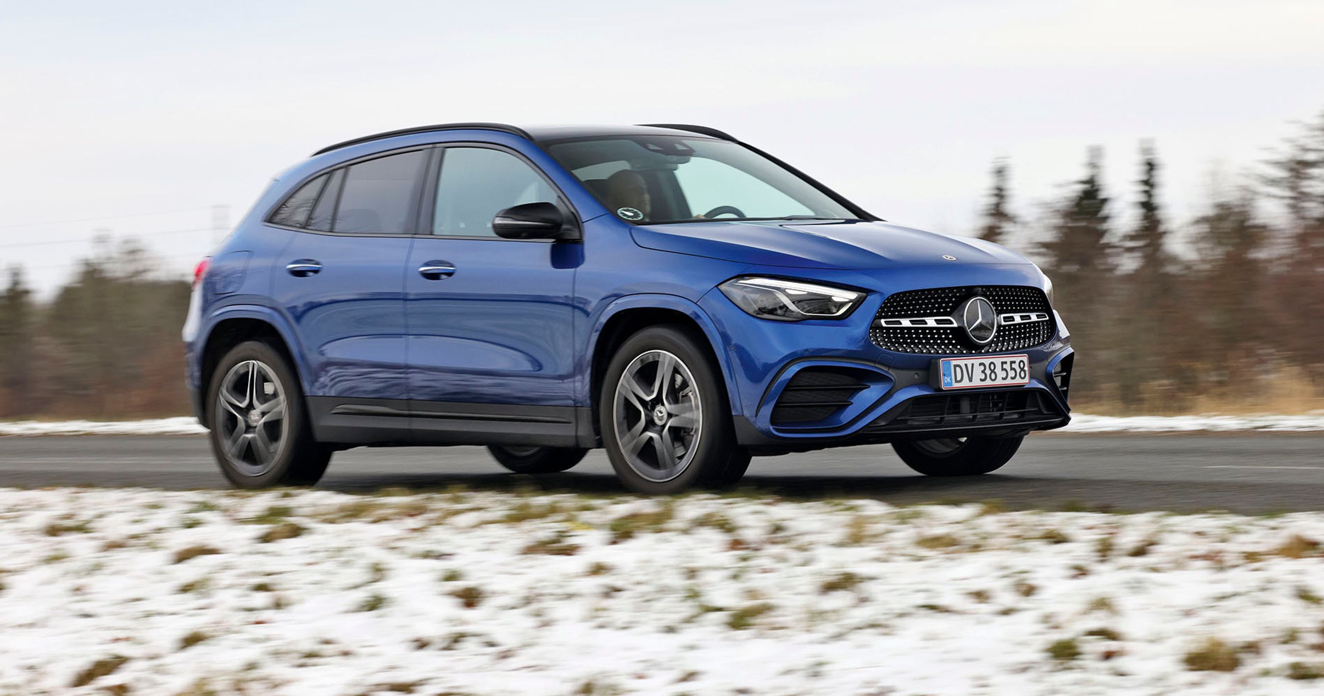 En blå Mercedes-Benz GLA 250 e kører på en landevej med sne i forgrunden og træer i baggrunden