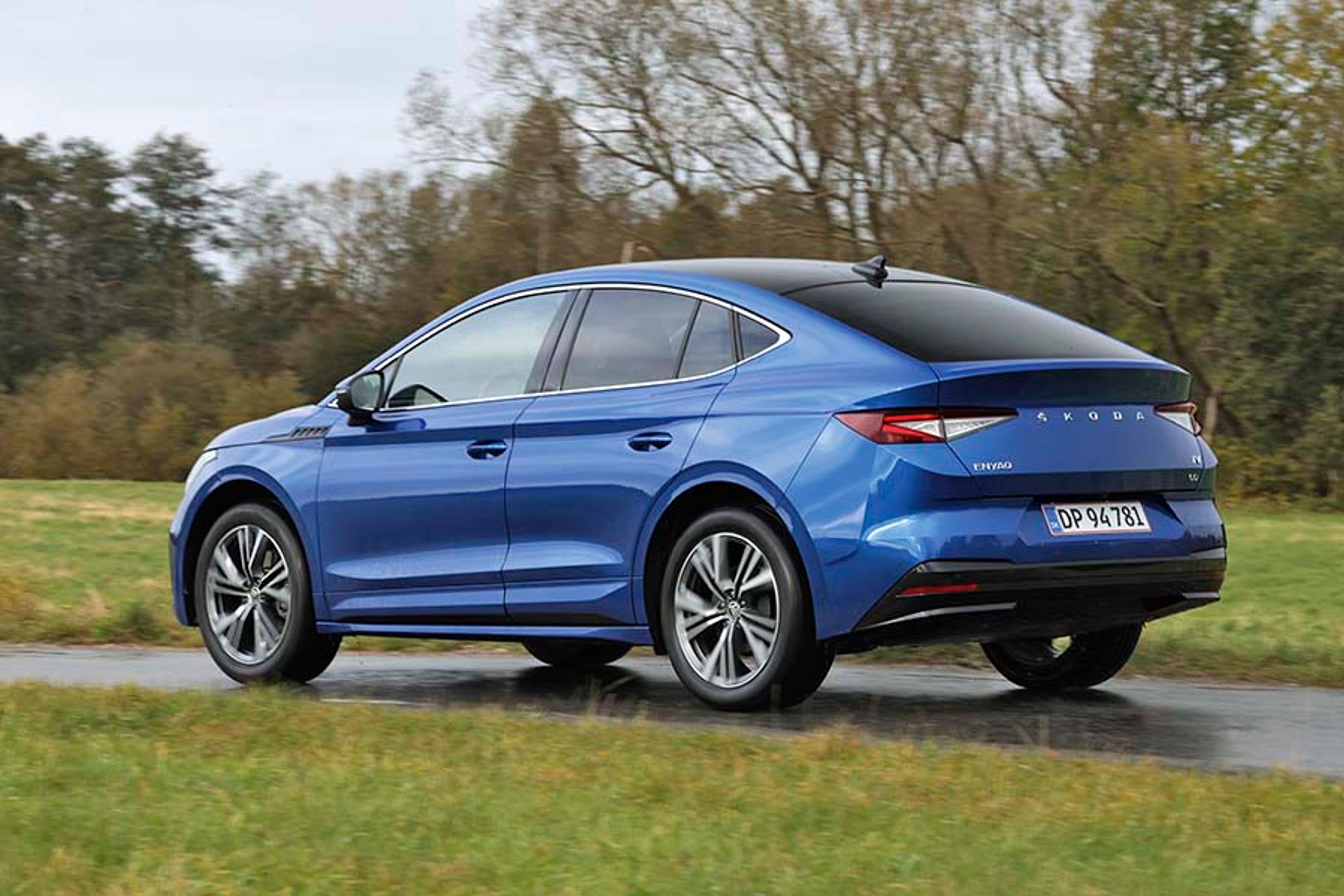 En blå Skoda Enyaq Coupé set bagfra, mens den kører på en landevej med græs i forgrunden
