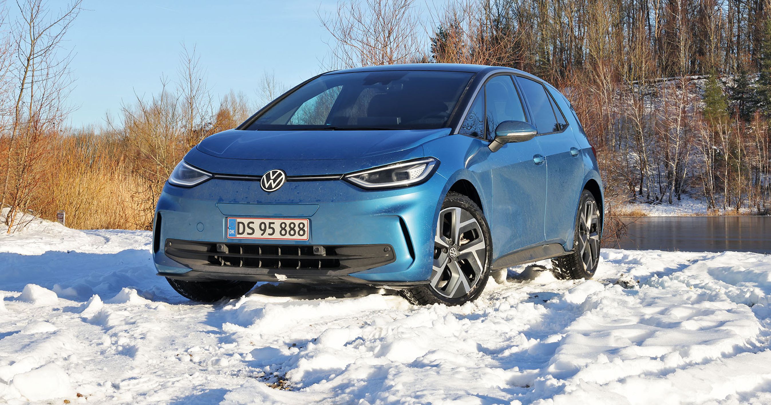 En blå VW ID.3 holder i sne med en skovsø i baggrunden