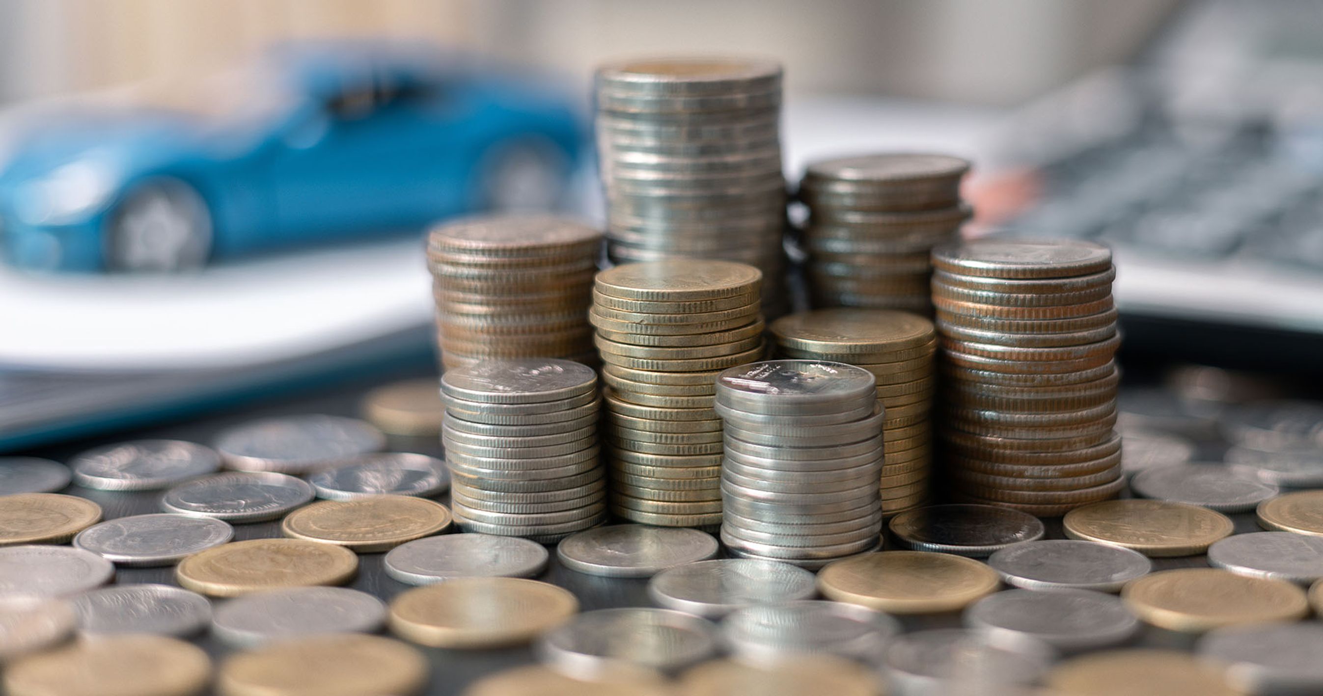 Mange mønter med en uskarp blå bil samt uskarp lommeregner i baggrunden. 