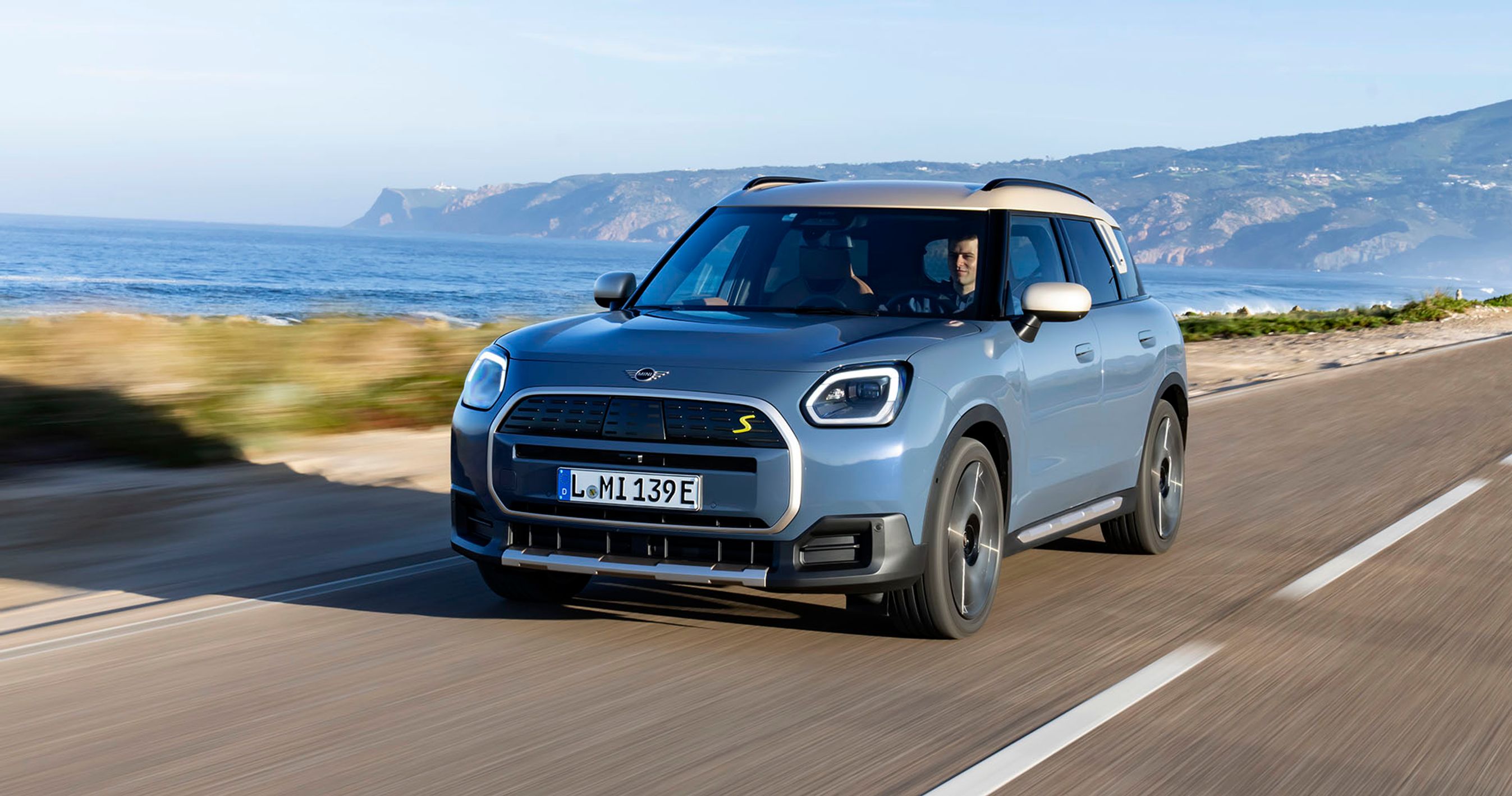 En blå Mini Countryman kører langs havet med bjerge i baggrunden