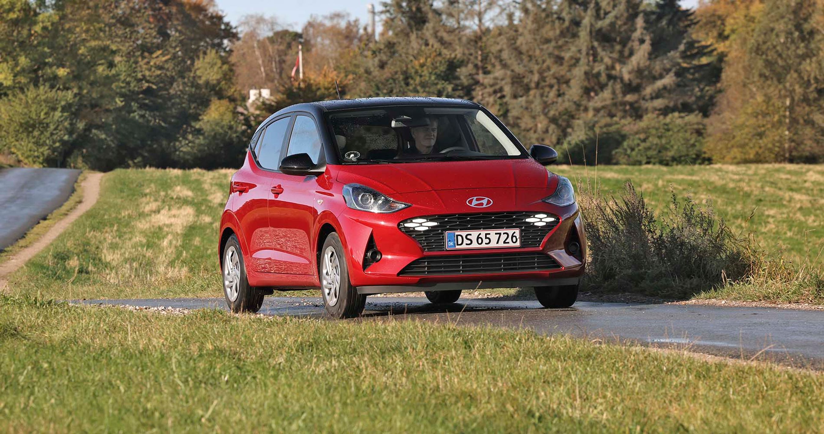 En rød Hyundai i10 kører på en landevej med træer i baggrunden