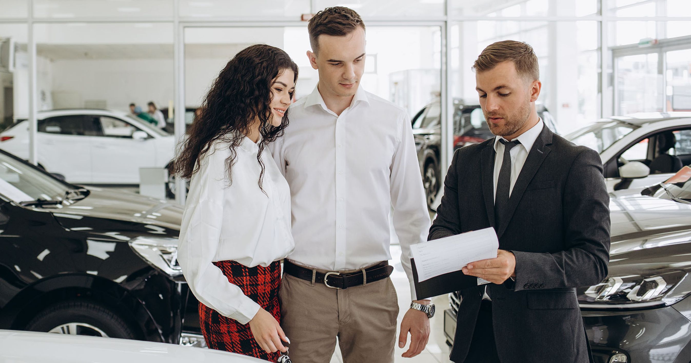 Tre personer står hos en bilforhandler og ser på et stykke papir