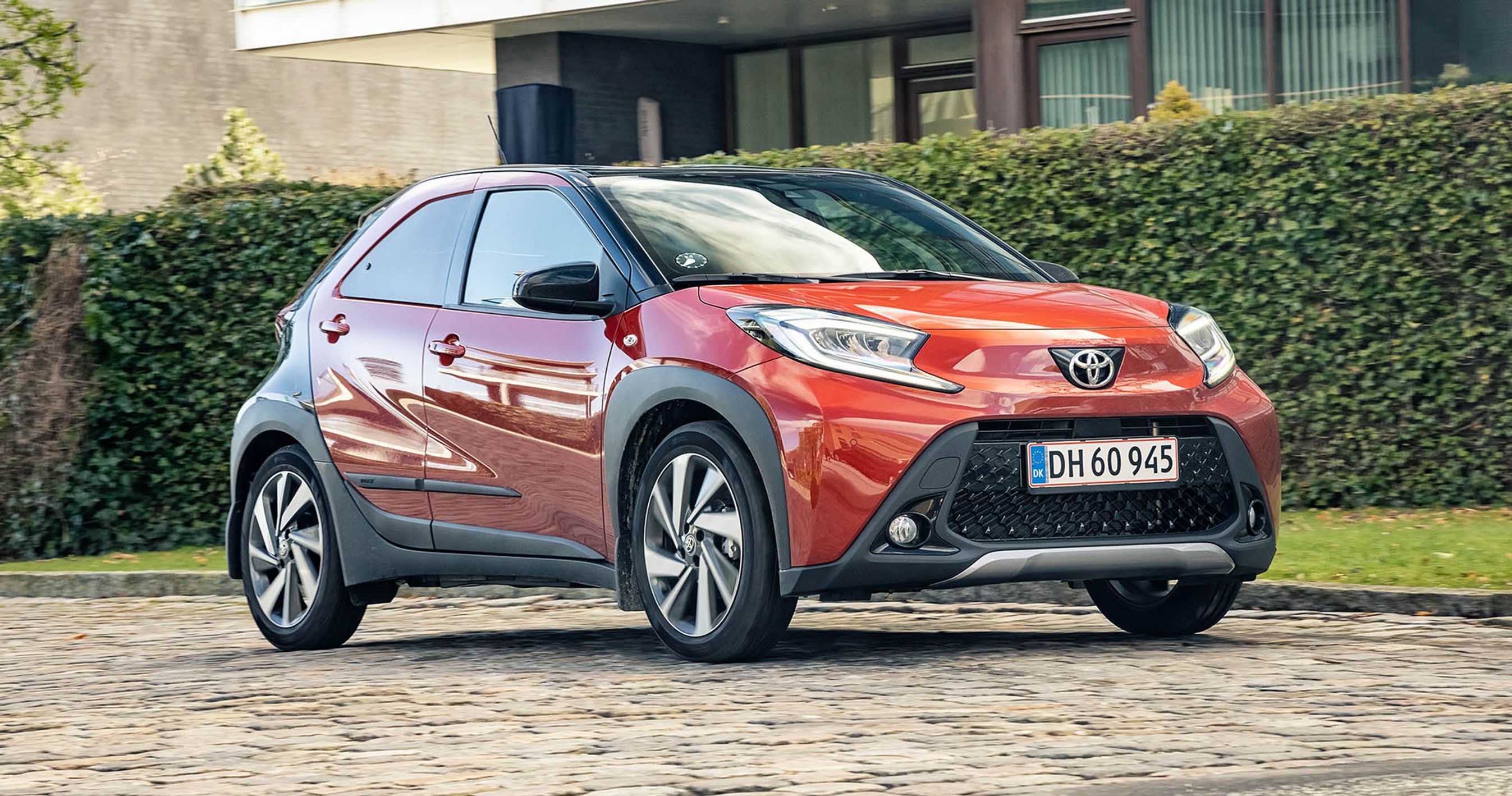 Orange Toyota Aygo X kører på brosten med grøn hæk i baggrunden.