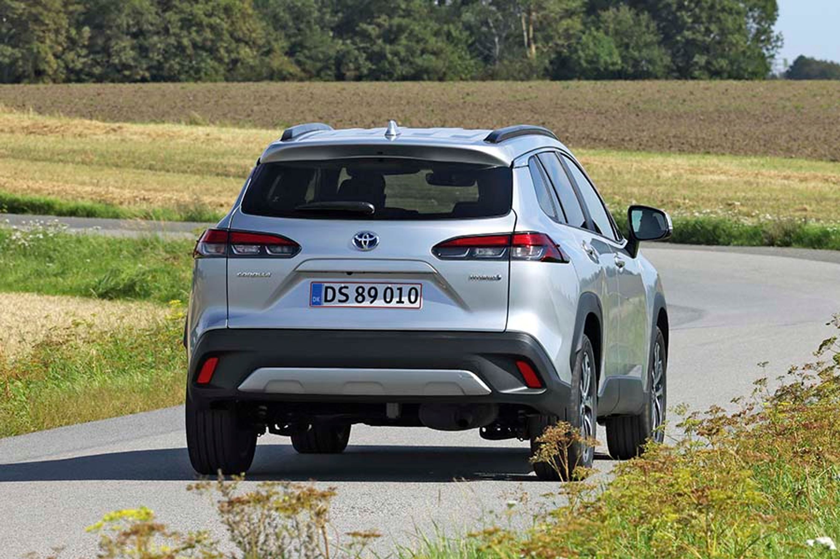 Toyota Corolla Cross set bagfra, mens den kører mod et sving på en landevej med marker rundtom.