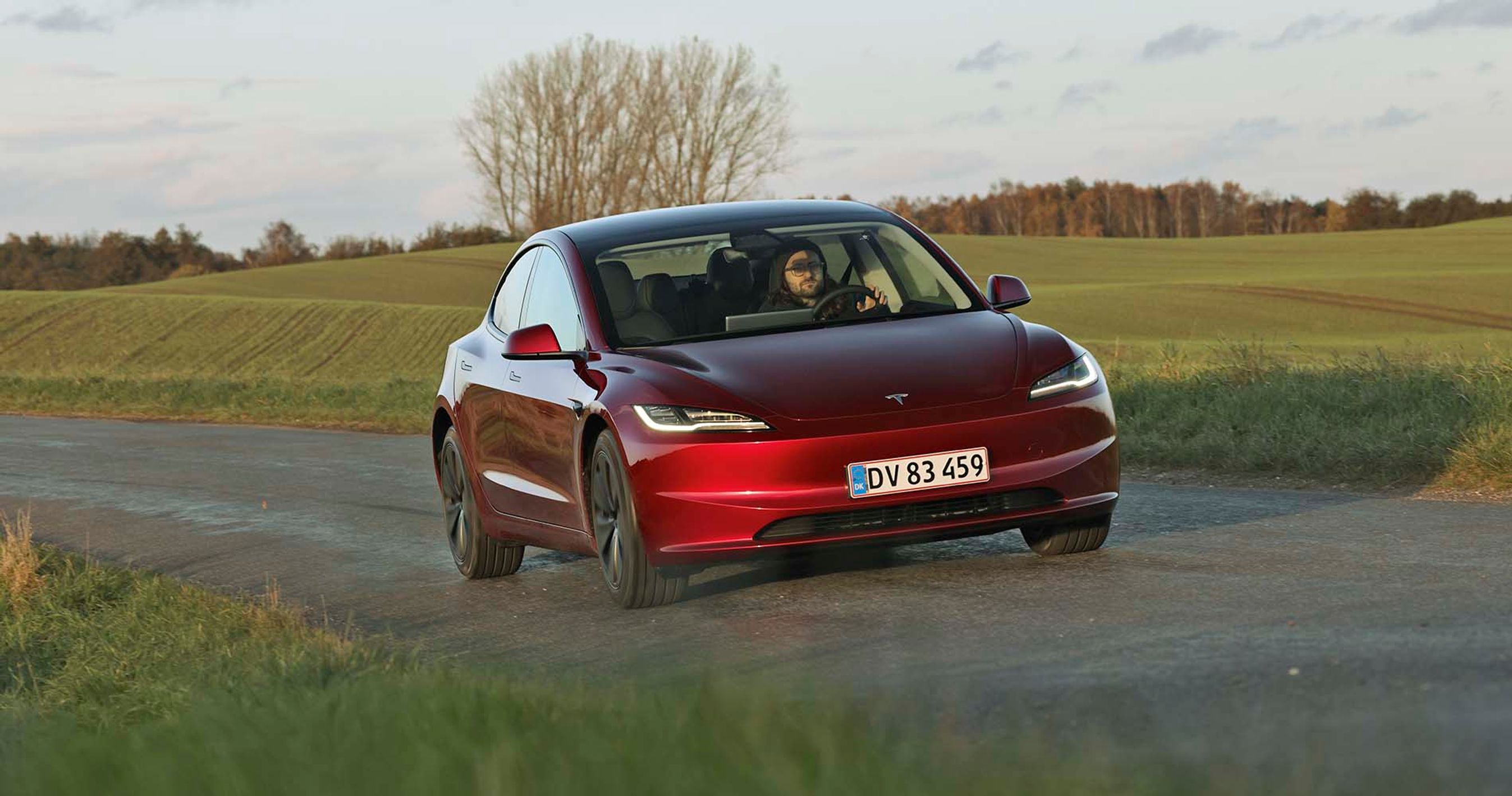En rød Tesla Model 3 kører på en landevej med græs og et træ i baggrunden.