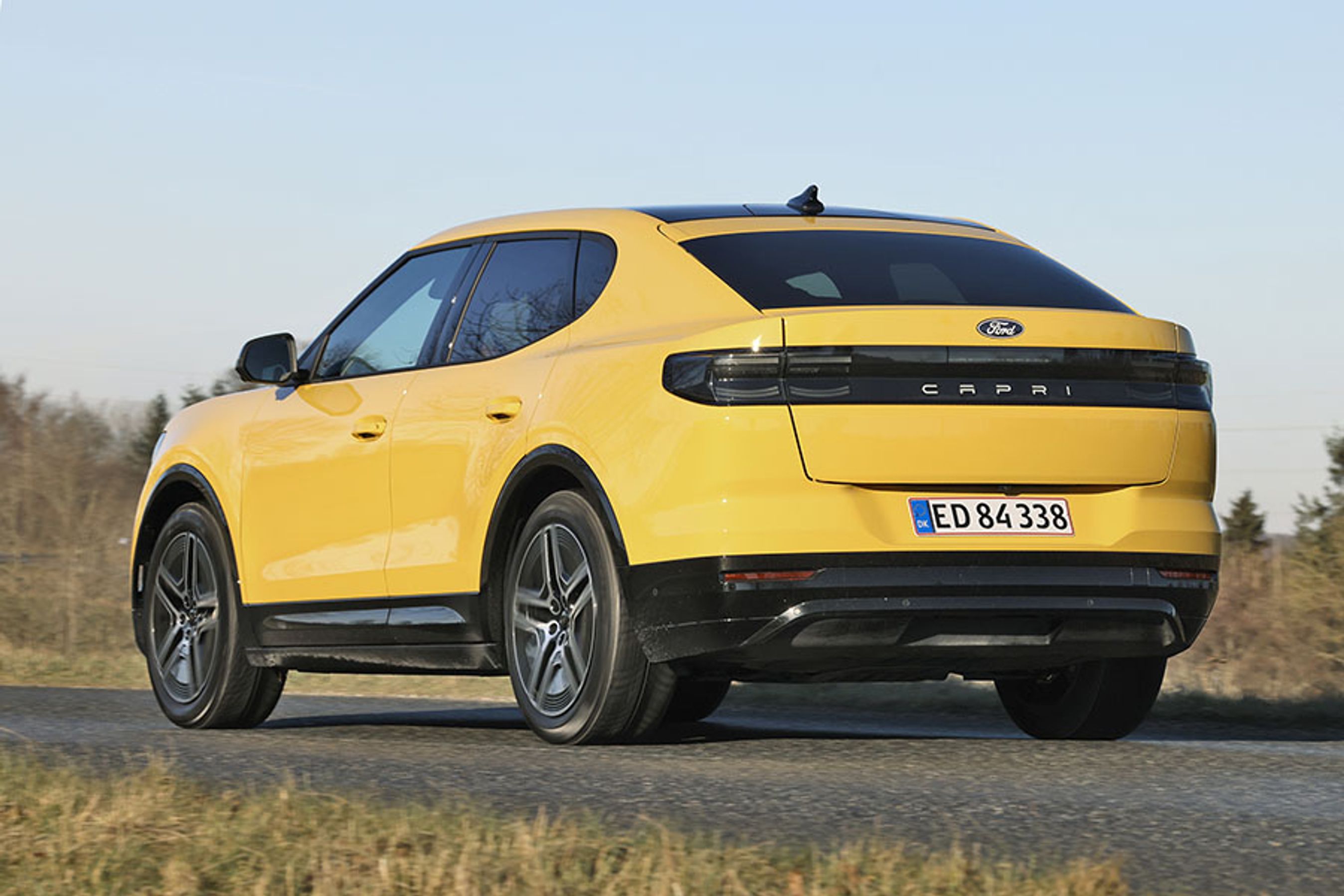 Gul Ford Capri set bagfra, mens den kører på en landevej med græs i forgrunden