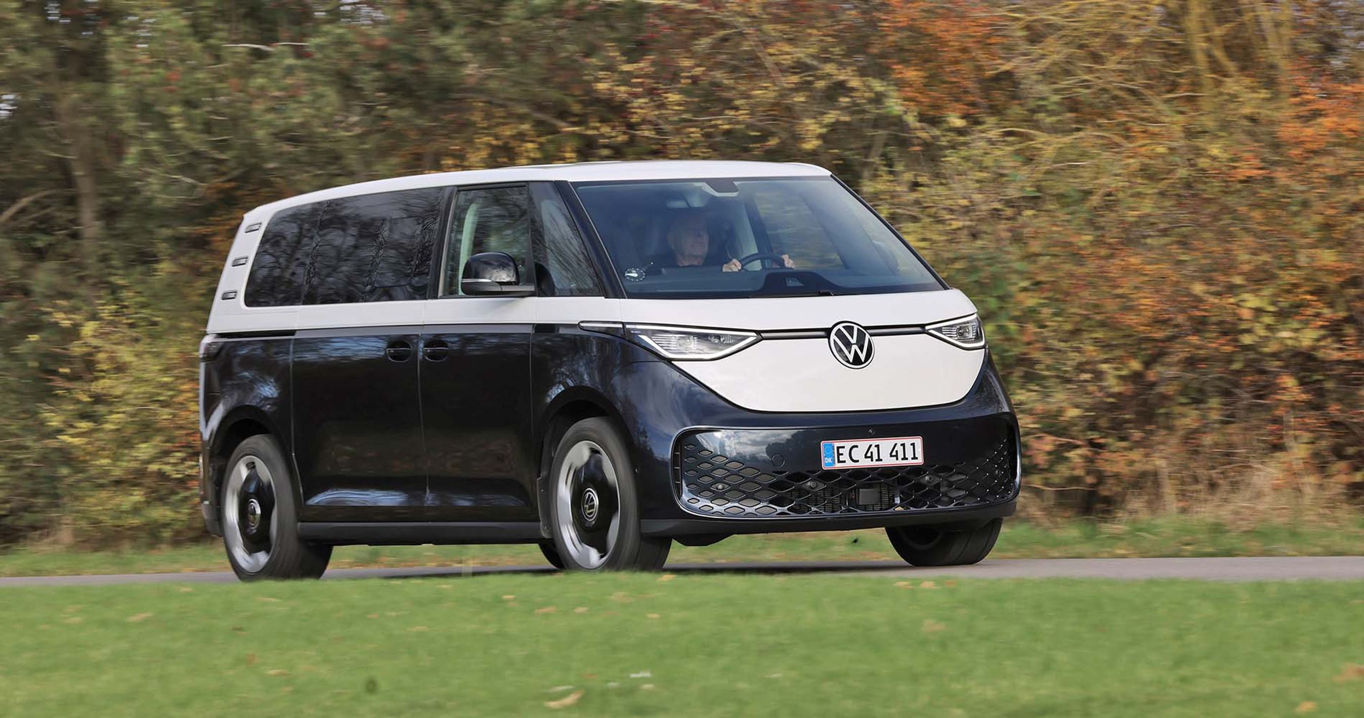 En blå og hvid Volkswagen ID. Buzz elektrisk minibus fotograferet under kørsel på en grøn græsmark med træer i baggrunden. 
