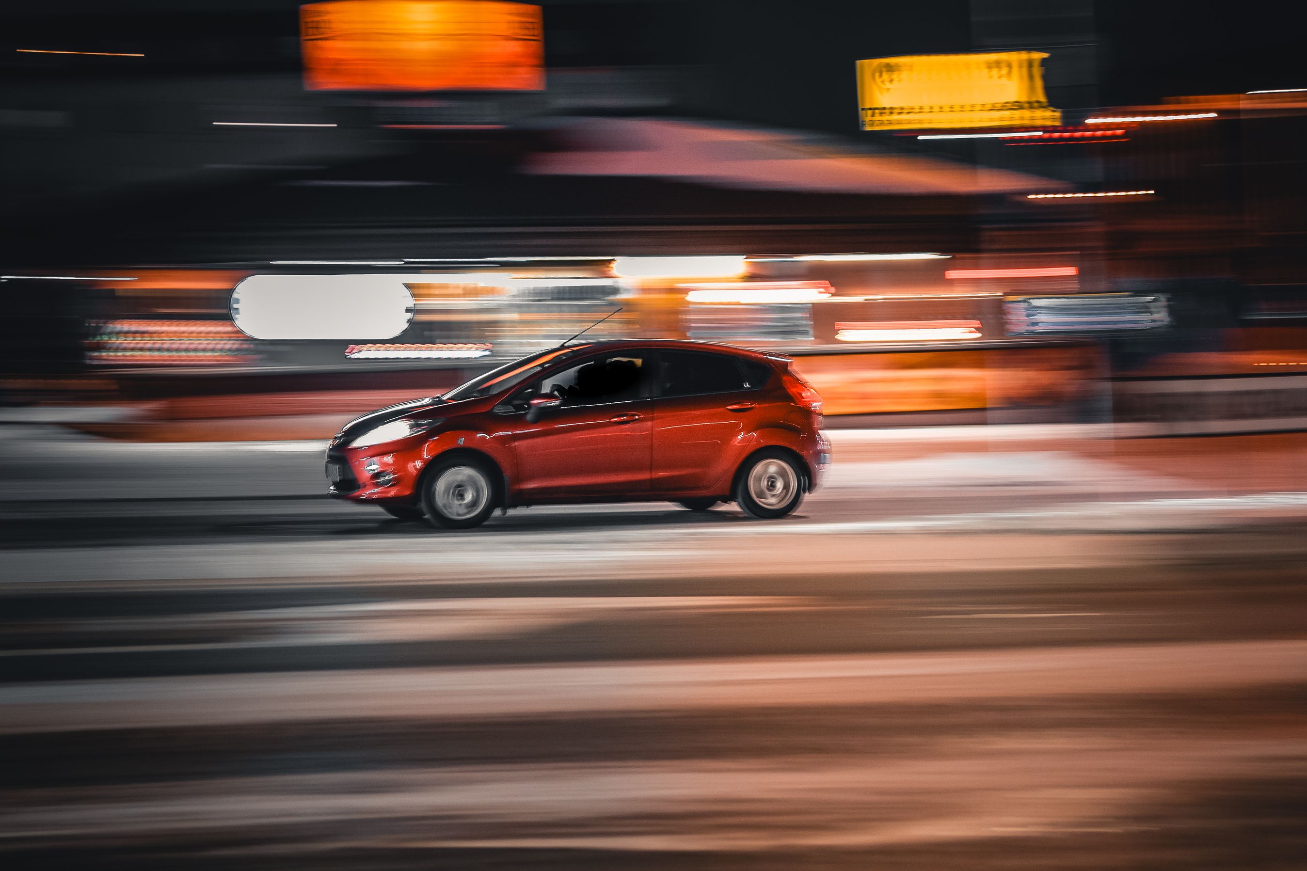 Rød bil kører hurtigt gennem en by om aftenen. Billedet er sløret pga. farten.
