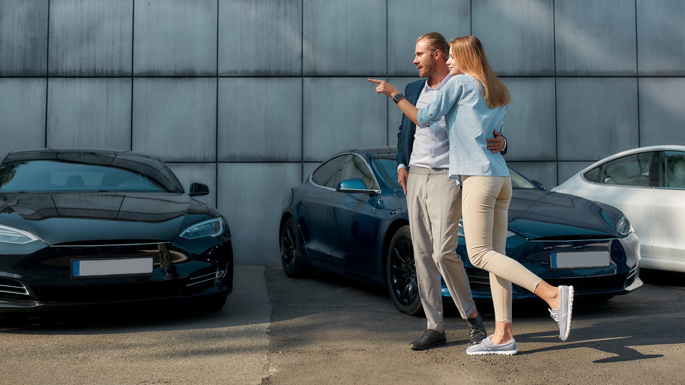 Et par står foran en række moderne elbiler på en parkeringsplads. Manden, der er klædt i en blazer og lyse bukser, peger mod noget i distancen, mens kvinden i en lyseblå skjorte og beige bukser smiler og læner sig ind mod ham med en arm omkring hans talje. I baggrunden ses en grå bygning med store facadeplader.