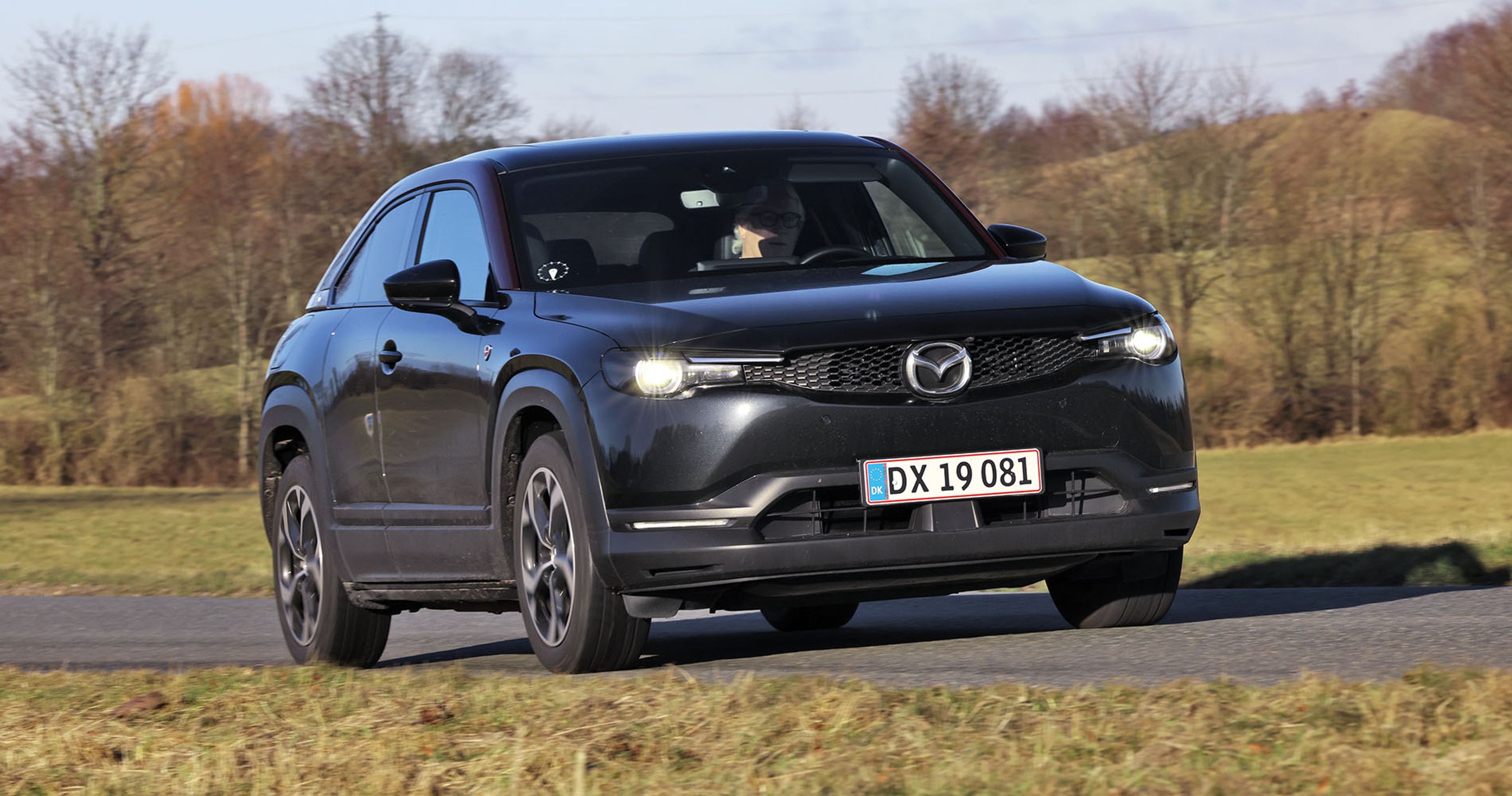 En sort Mazda MX-30 R-EV kører på en landevej med træer og græs i baggruden