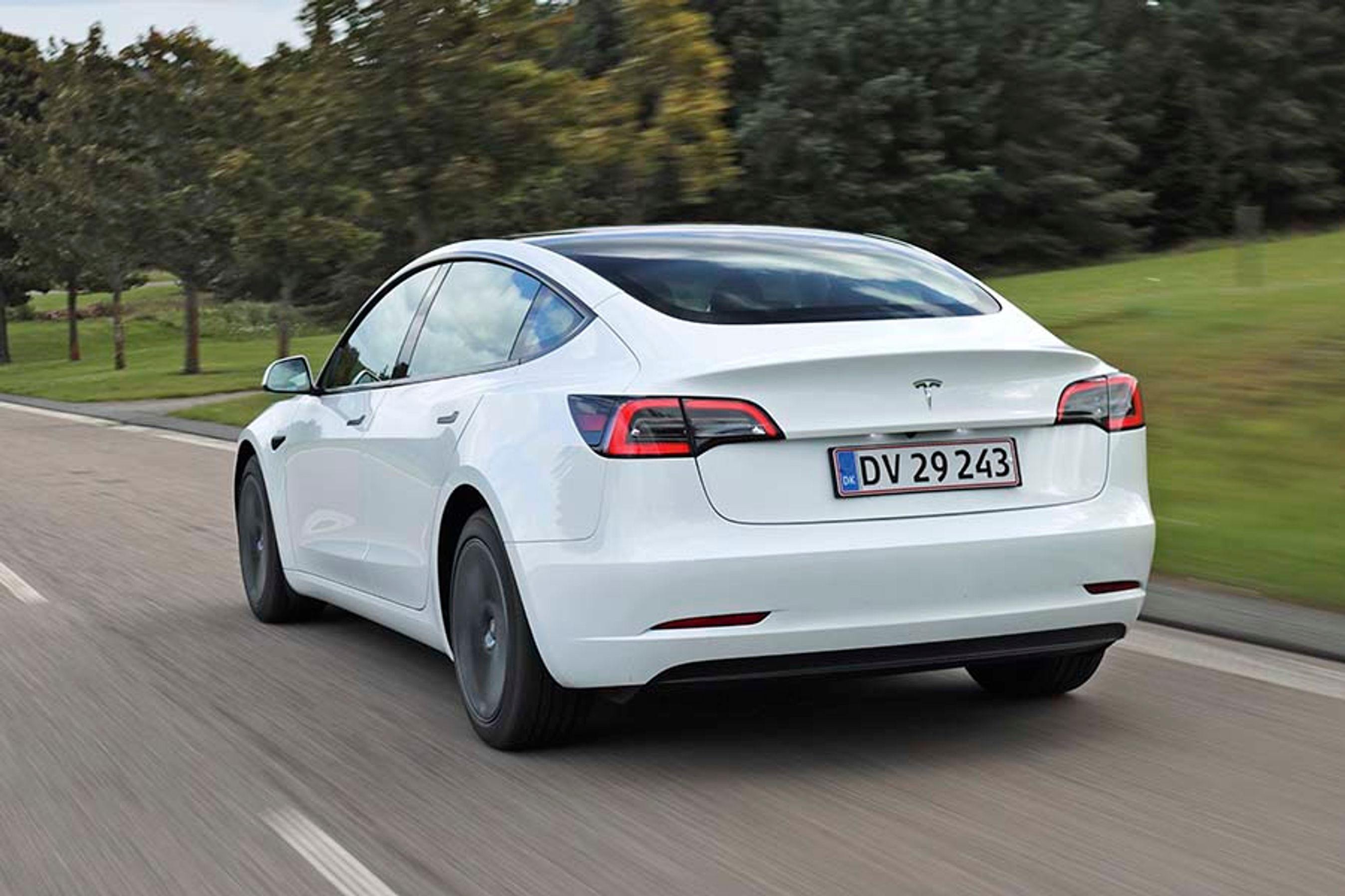 En hvis Tesla Model 3 set bagfra kører ad en landevej med træer i baggrunden.