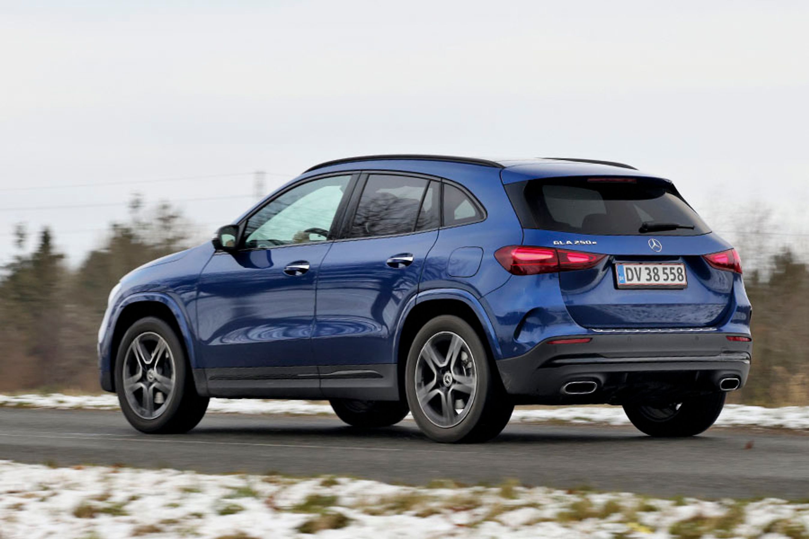 En blå Mercedes-Benz GLA 250 e set bagfra, mens den kører på en landevej med sne i forgrunden og træer i baggrunden.