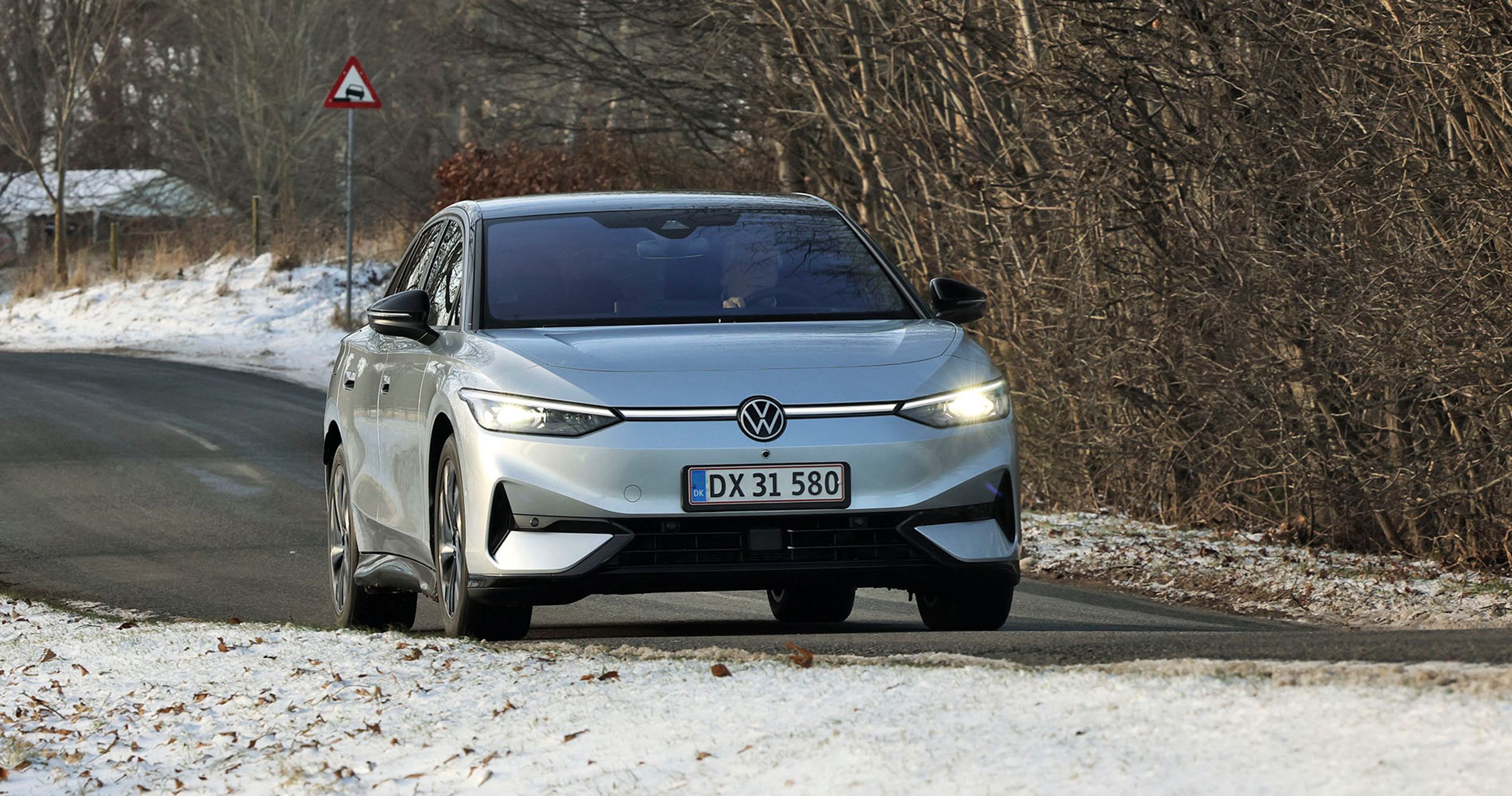 En sølvgrå VW ID.7 kører på en landevej med sne i forgrunden og træer i baggrunden