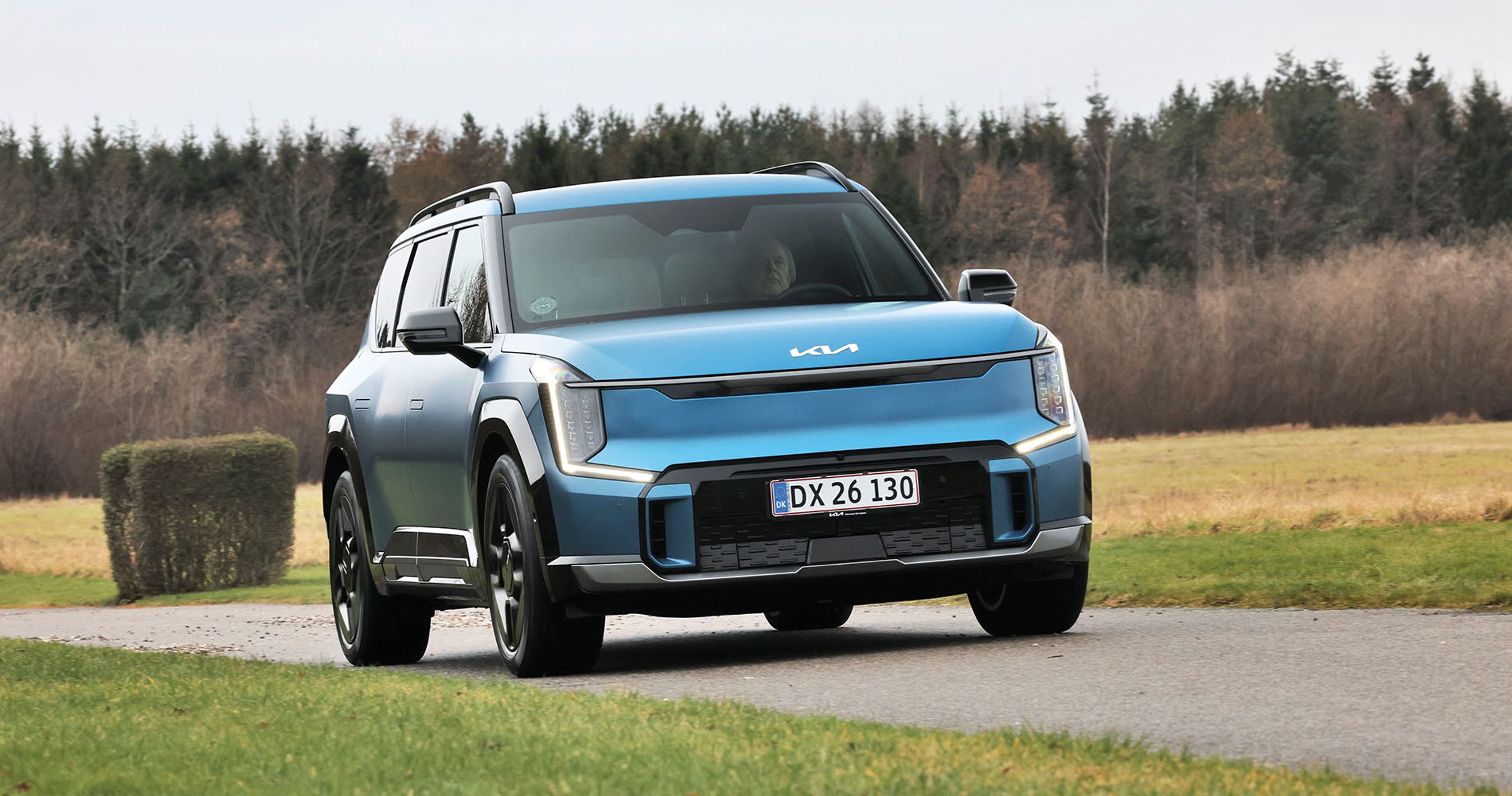 En blå Kia EV9 kører på en landevej med græs i baggrudnen