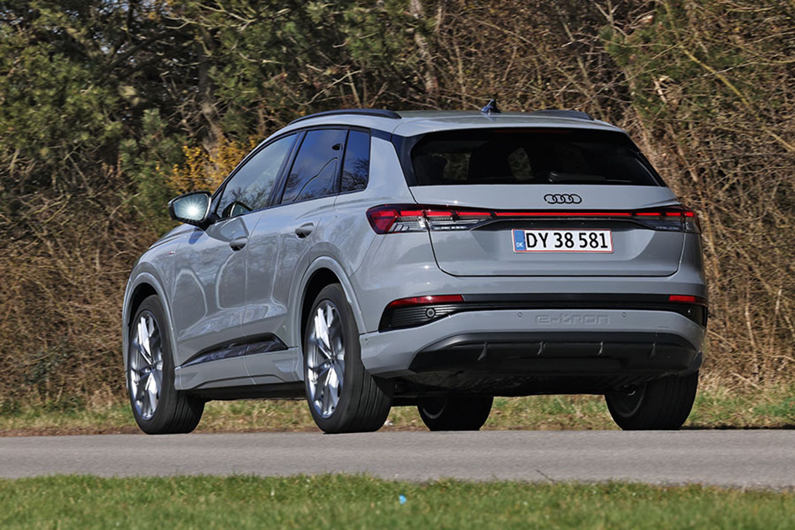 En grå Audi Q4 e-tron set bagfra kører på en vej med træer i baggrunden