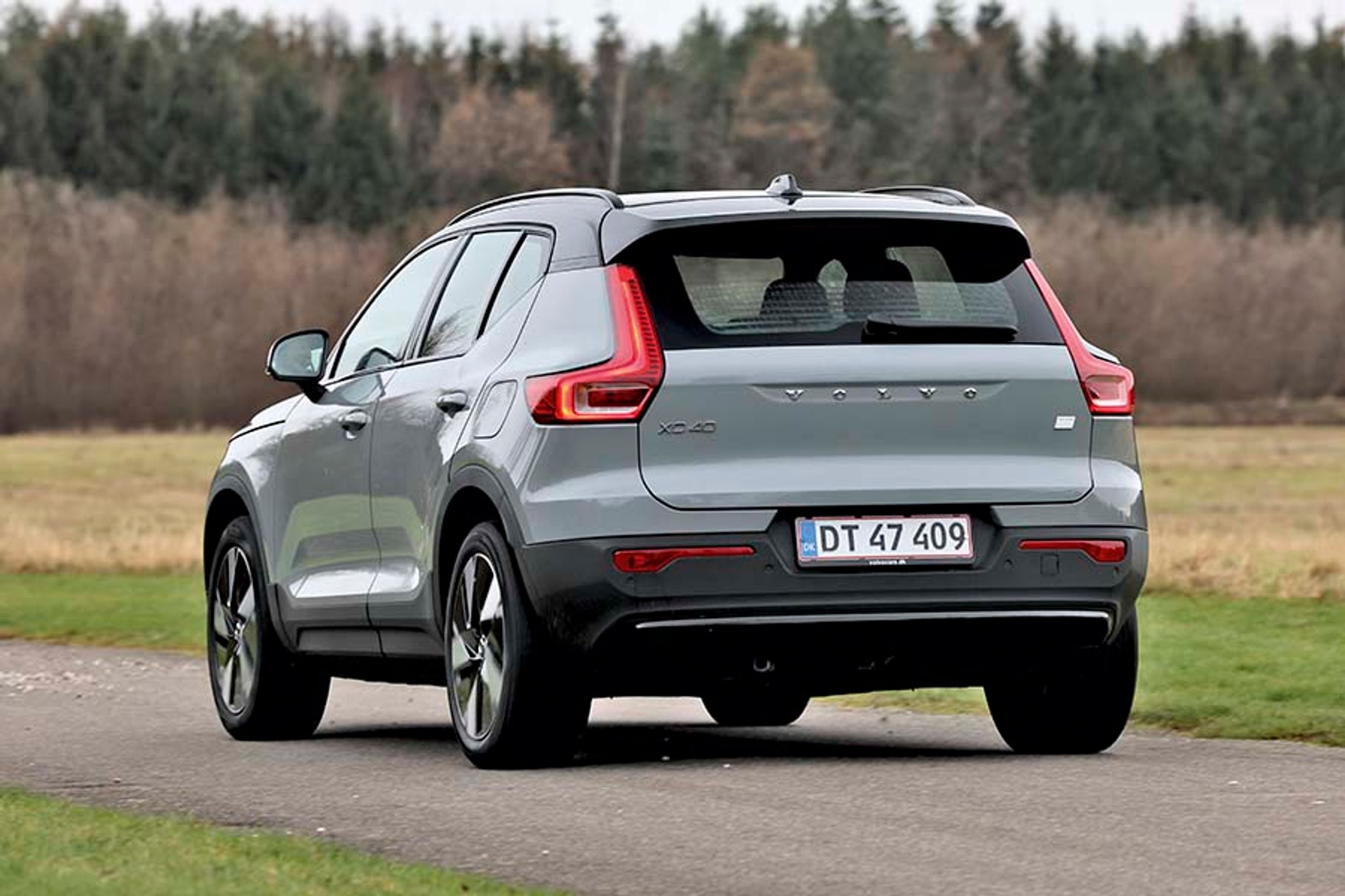 En grå Volvo XC40 elbil set bagfra, mens den kører på en landevej med træer i baggrunden