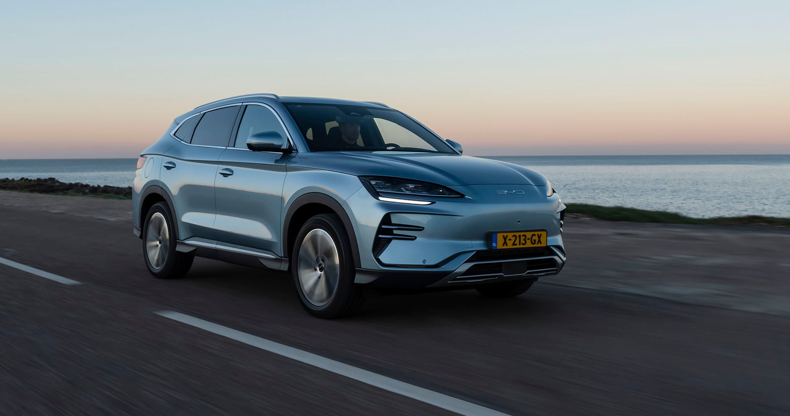 En sølvgrå BYD Seal U kører langs havet ved solnedgang.